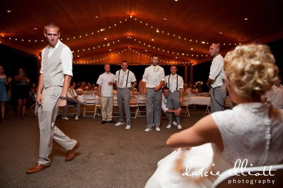A group of men dressed in formal attire are standing in a line during a wedding reception. An elderly woman in a white dress is sitting in the foreground, facing the men. The venue has a wooden ceiling with string lights, and there's a table with flo