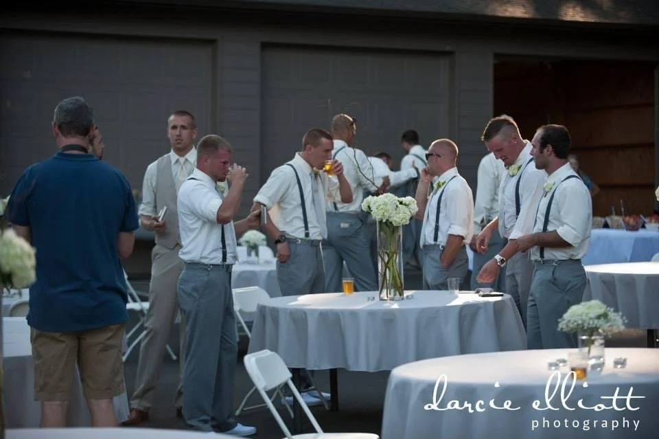 Group of men in formal attire gathered around a table at an outdoor event, some drinking and chatting, with white floral centerpieces on round tables