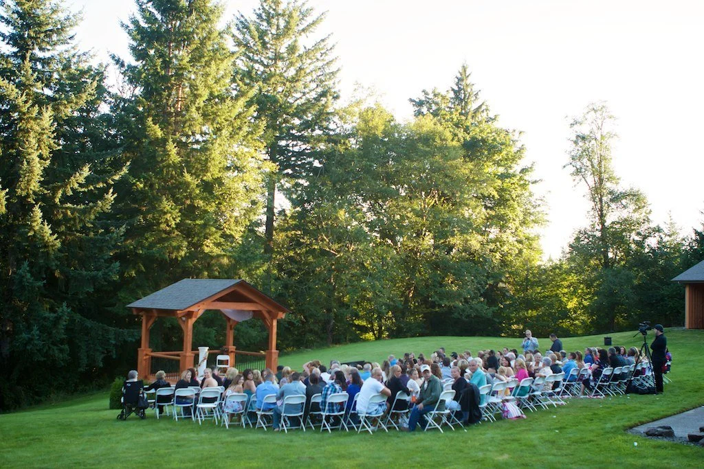 Outdoor wedding ceremony with seated guests on chairs facing a wooden altar under a small pavilion in a grassy area, surrounded by tall green trees.
