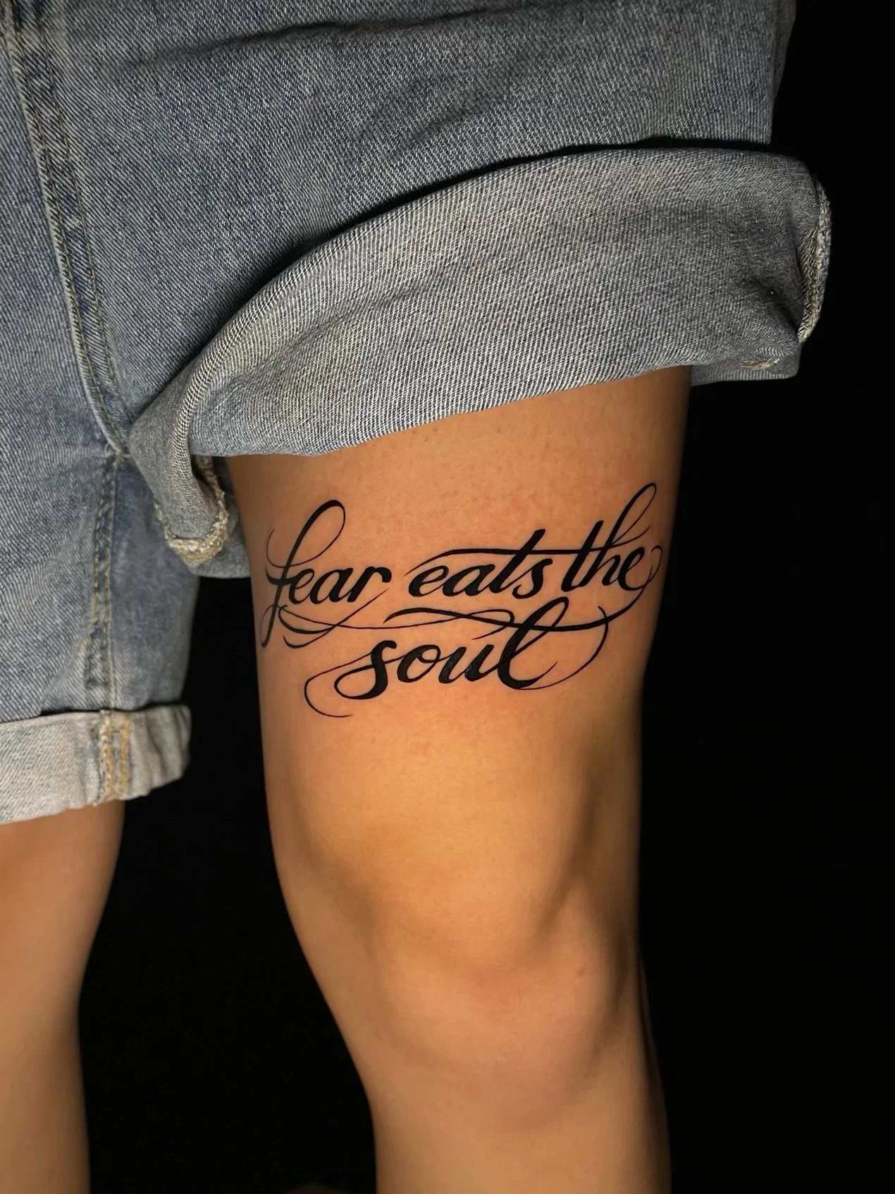 Elegant “Fear Eats the Soul” lettering tattoo on the upper thigh, featuring bold black calligraphy, flowing cursive script, and intricate decorative flourishes—tattoo by Hayden Herndon at Cloak Collective.