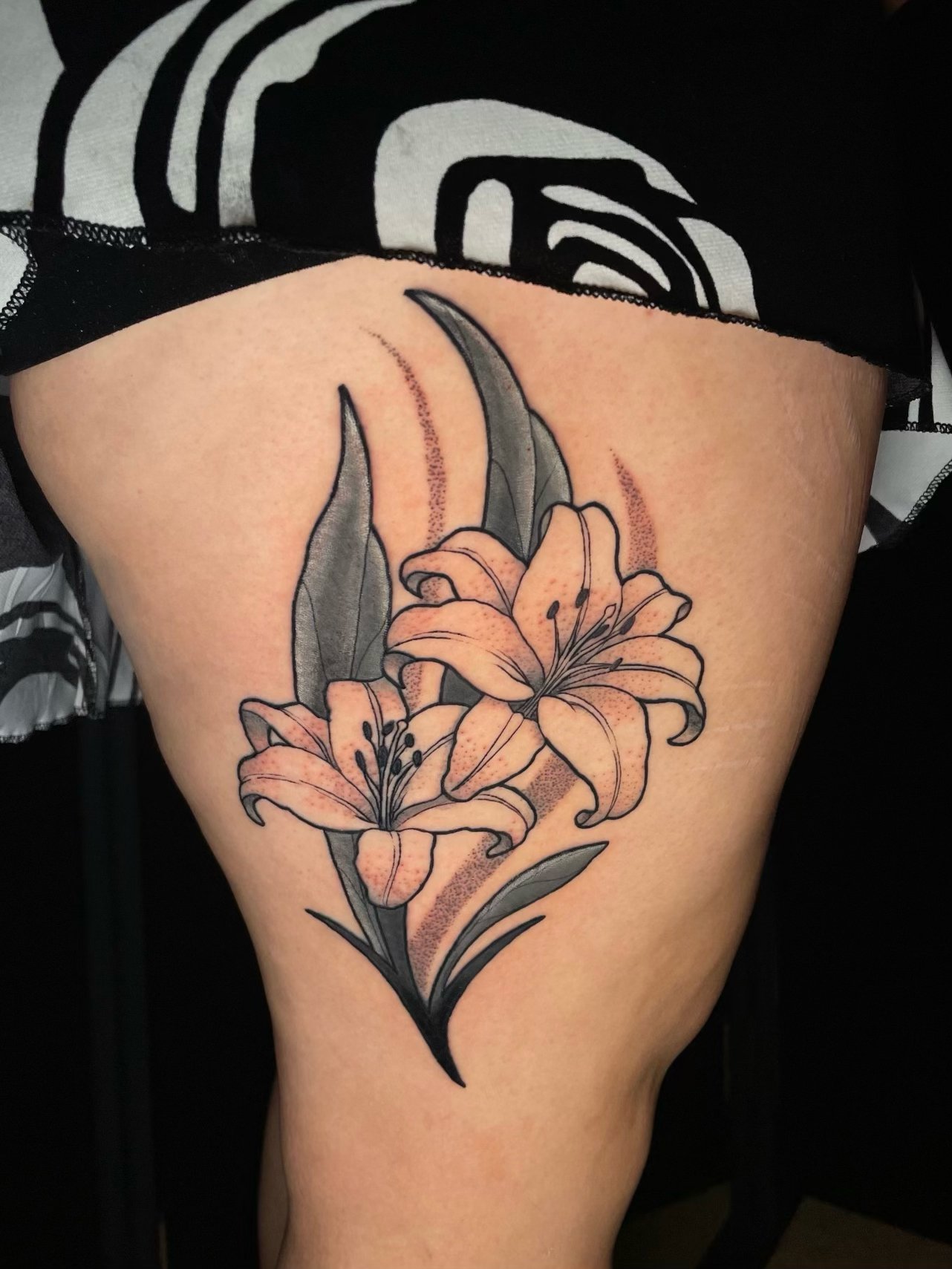 Elegant floral thigh tattoo featuring two blooming lilies with delicate black linework, soft peach-pink petals, detailed dotwork shading, and bold black and gray leaves—tattoo by Hayden Herndon at Cloak Collective.