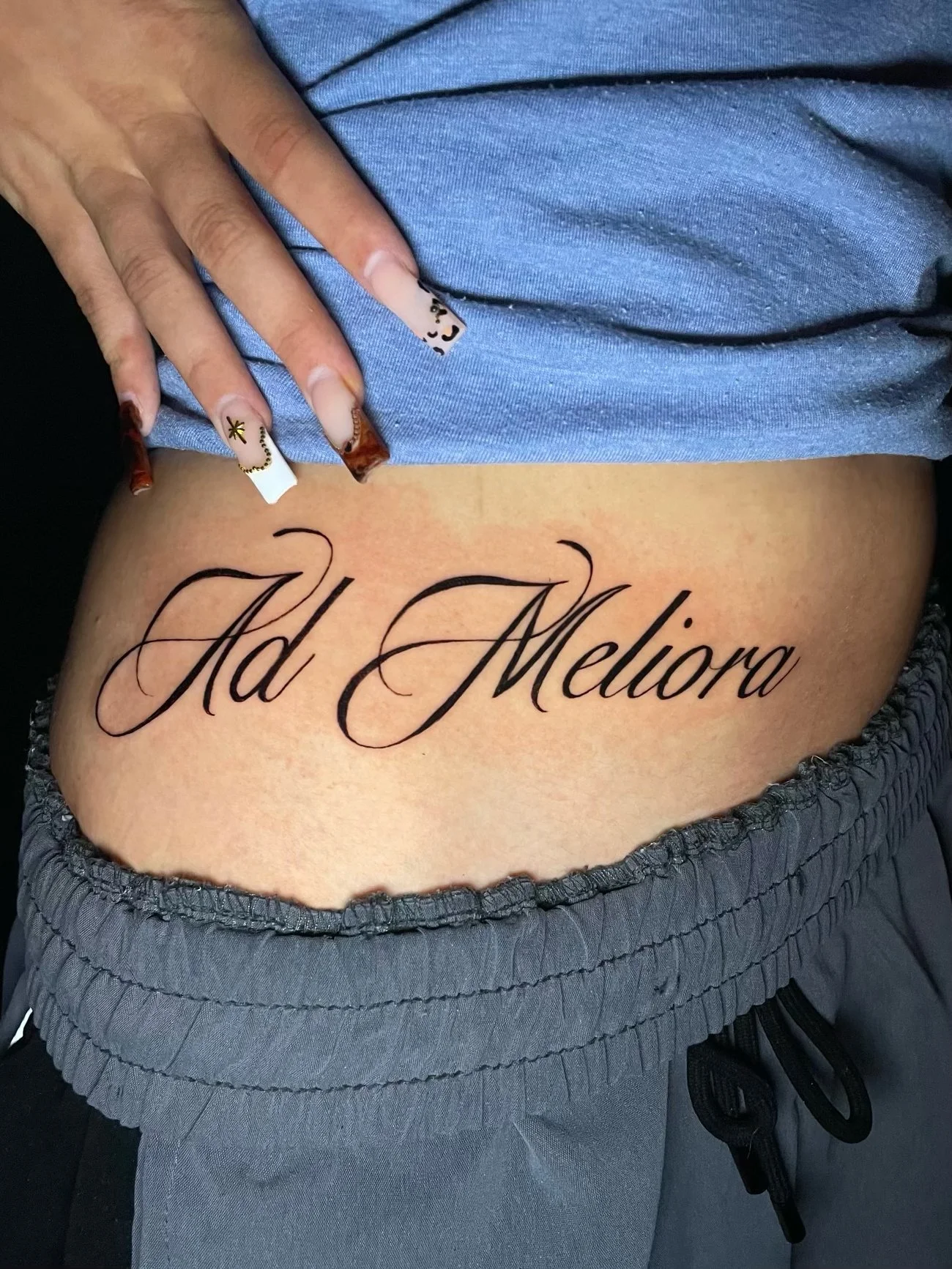 Elegant “Ad Meliora” script tattoo across the lower abdomen featuring flowing black calligraphy, delicate flourishes, and refined lettering symbolizing the pursuit of better things—tattoo by Hayden Herndon at Cloak Collective.