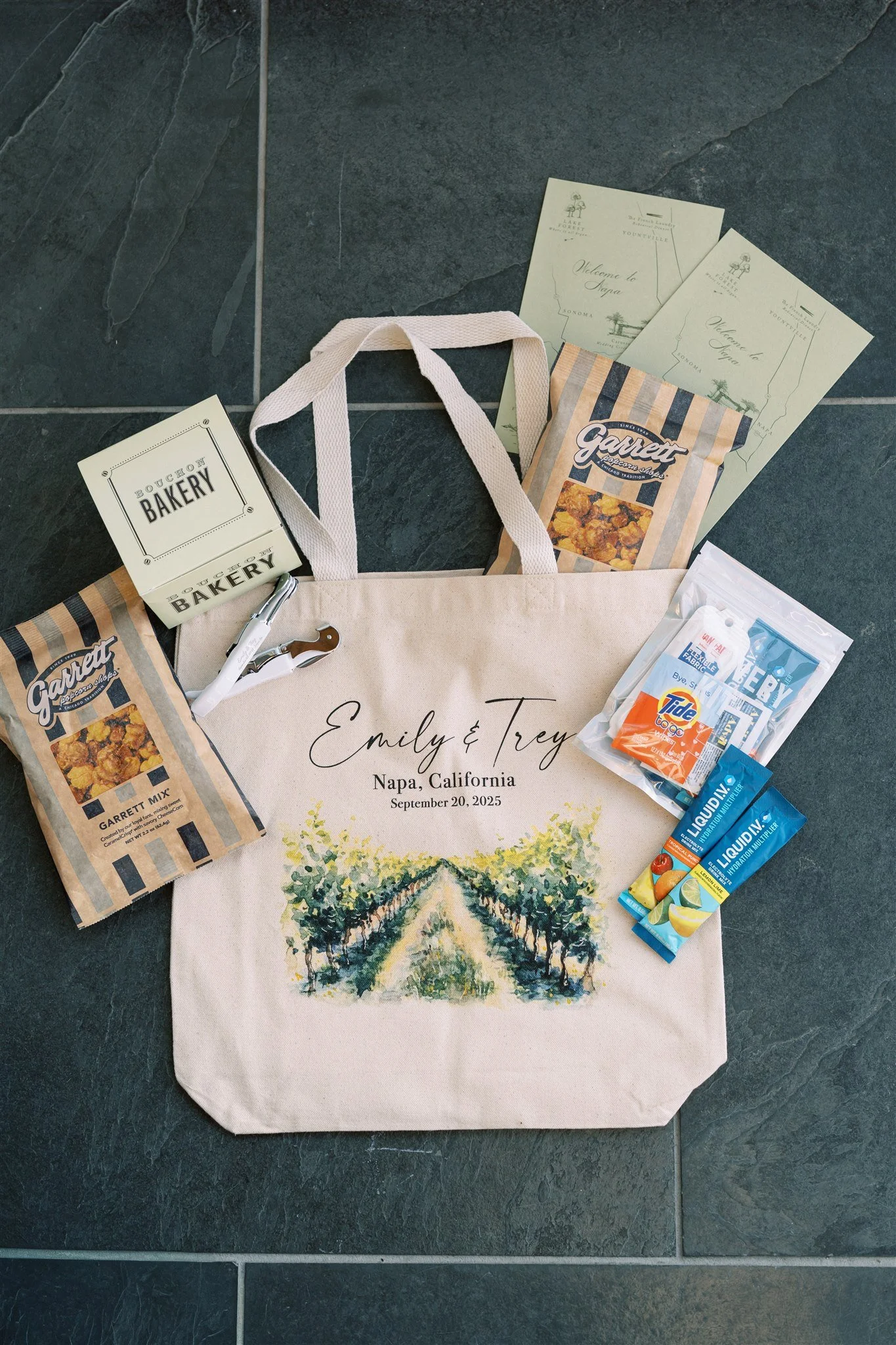A tote bag featuring a landscape with vinyards and trees, with the text 'Emily & Trey Napa, California September 20, 2025' printed on it. Surrounding the bag are two packets of Garrett popcorn, a small box of bakery items, and several flyers or brochures.