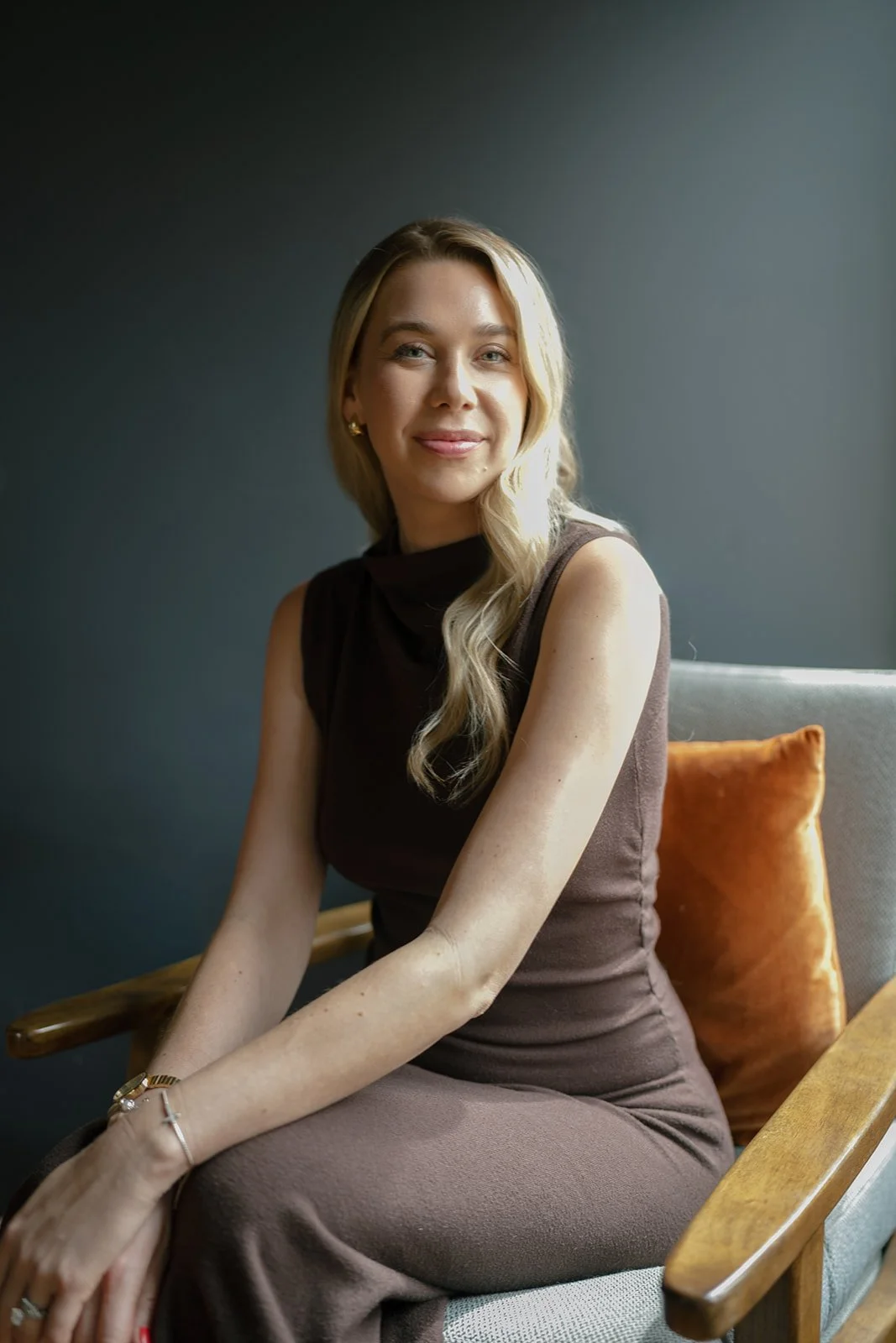 A woman with long blonde hair in a brown sleeveless dress sitting on a wooden chair with a cushion, smiling at the camera.