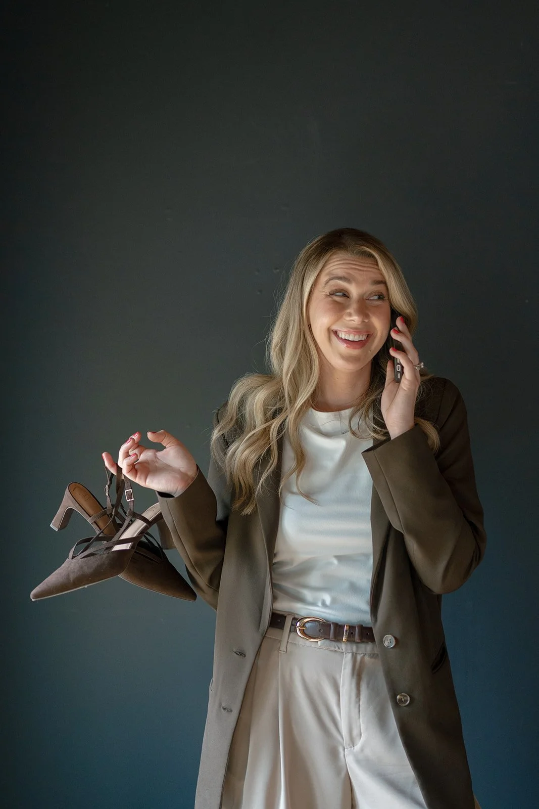 Lexi is smiling while talking on her cell phone, holding a pair of high-heeled shoes in her other hand, wearing a white blouse, beige pants, and a brown blazer, standing against a dark gray background.