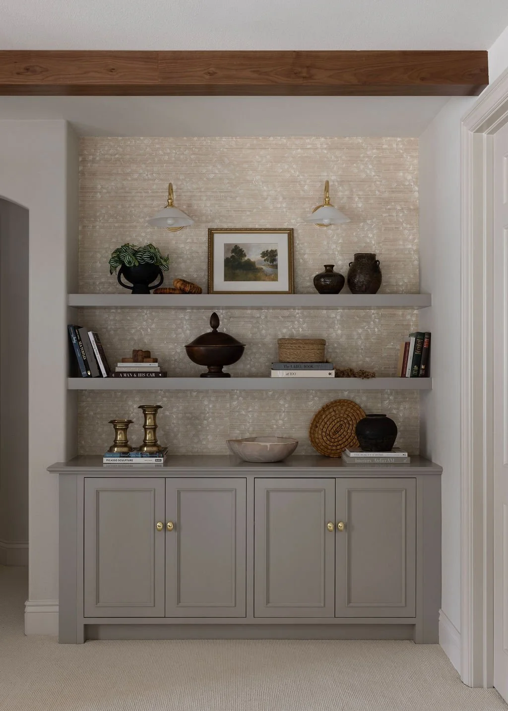 A built-in bookshelf unit with three shelves and a gray cabinet base, decorated with vases, books, and artwork, against a textured beige wall with wall sconces.