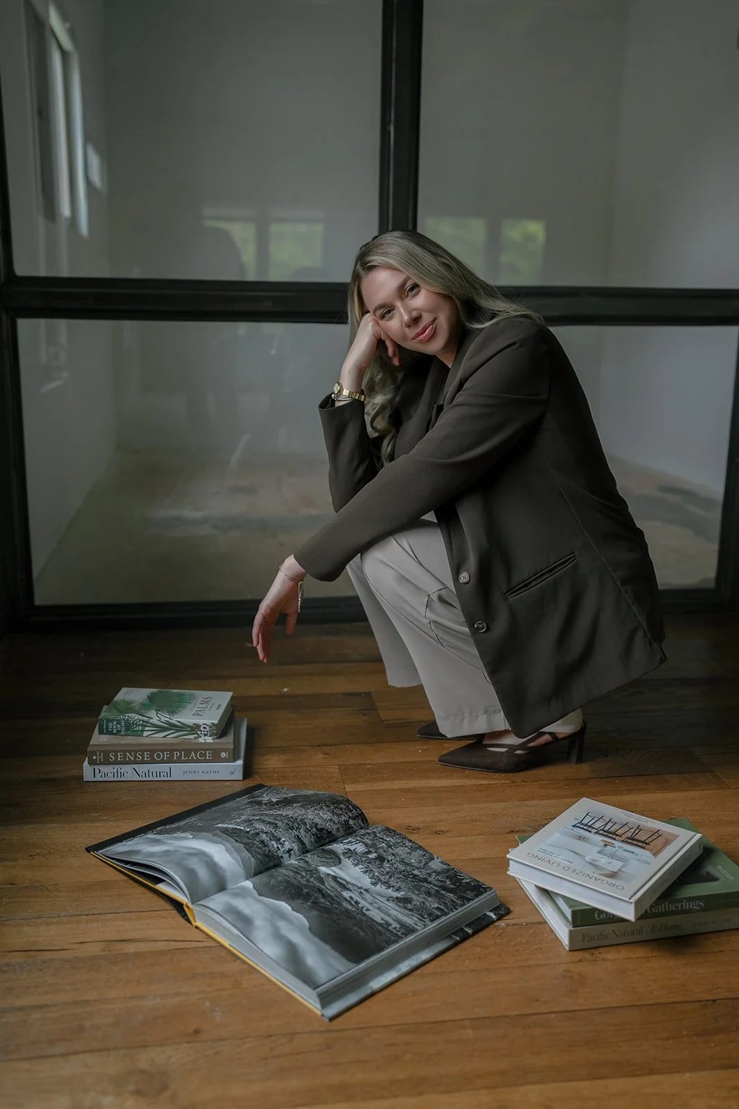 Lexi, owner, in a brown blazer and beige pants crouches on a wooden floor next to a stack of books and an open photo book. She is indoors near large windows, with a contemplative expression.