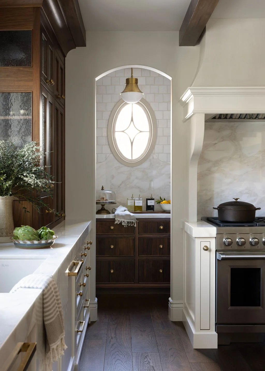 A kitchen with white countertops, wooden cabinets, and a stove with a pot. There are decorative jars, a bowl of lemons, and a plant on the countertops. An archway reveals a small area with dark cabinetry, a window with decorative trim, and a hanging gold and white pendant light.