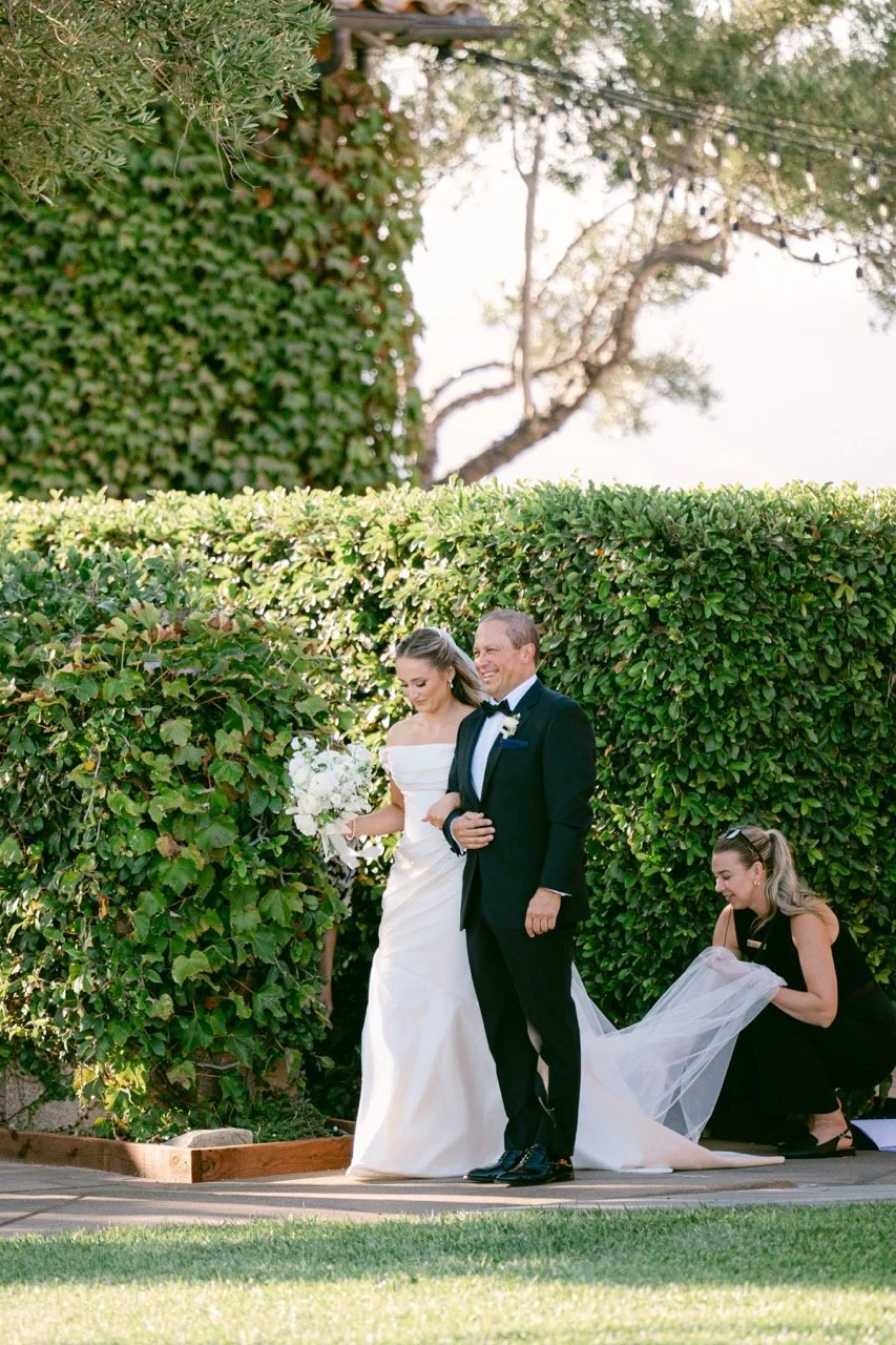 A bride in a white wedding gown holding a bouquet next to a smiling man in a tuxedo, with a woman adjusting her gown behind them outdoors, surrounded by green hedges and a tree.