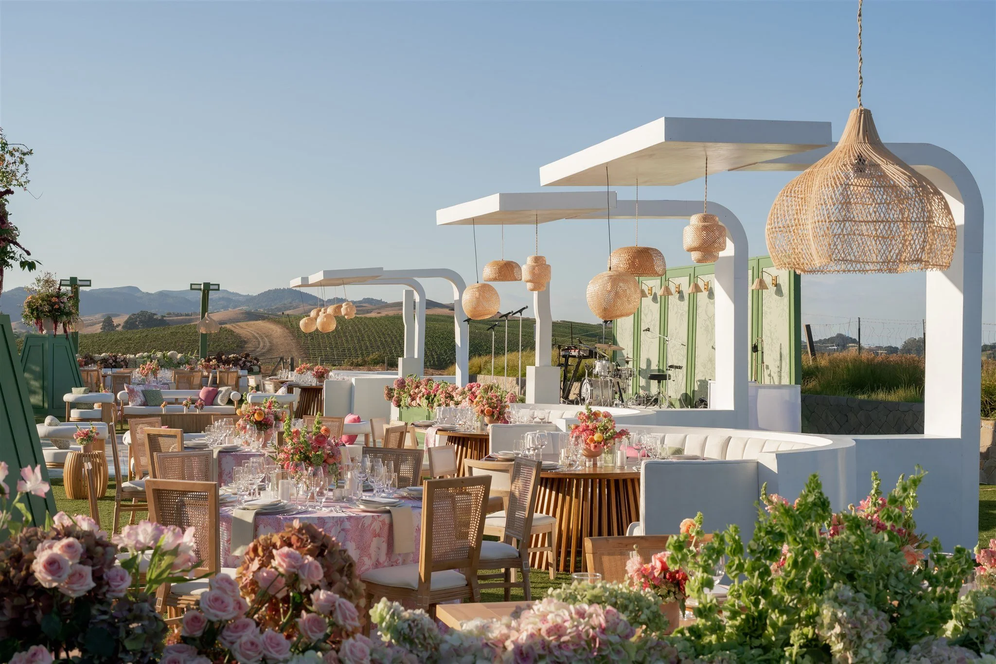 Outdoor wedding reception setup with tables decorated with flowers, surrounded by greenery and scenic mountains in the background. Hanging woven lamps add decor, with a view of a clear blue sky.