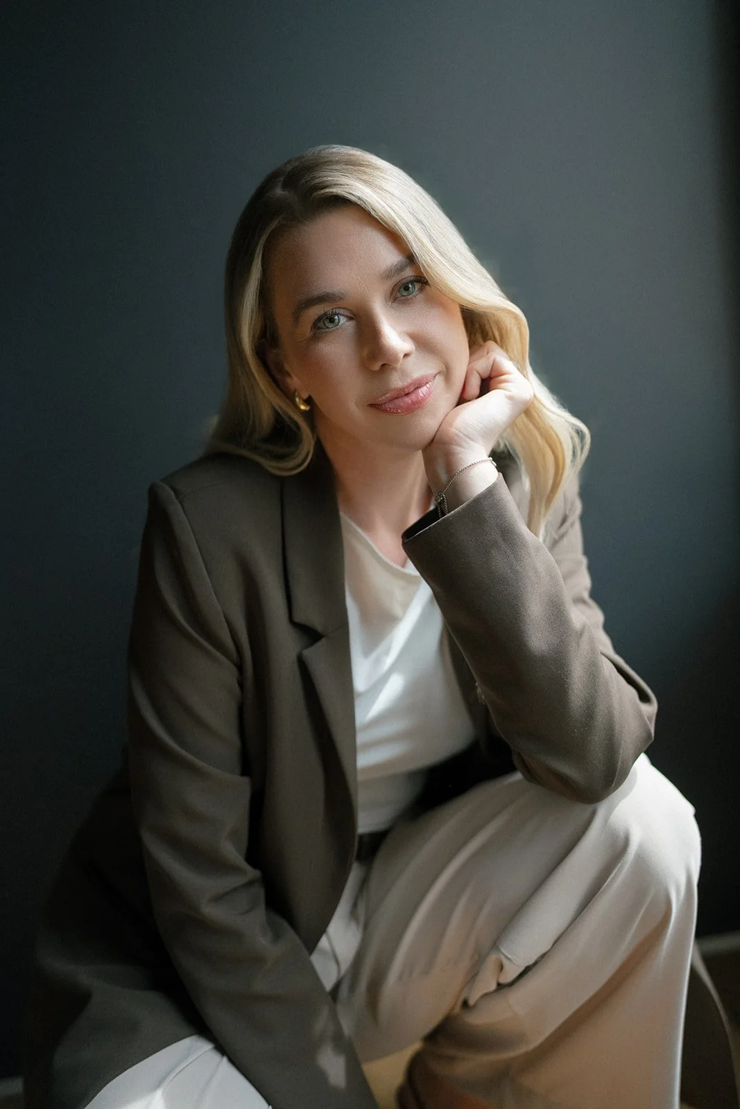 A portrait of a woman with blonde hair, sitting against a dark background, resting her chin on her hand, wearing a brown blazer, white shirt, and beige pants.