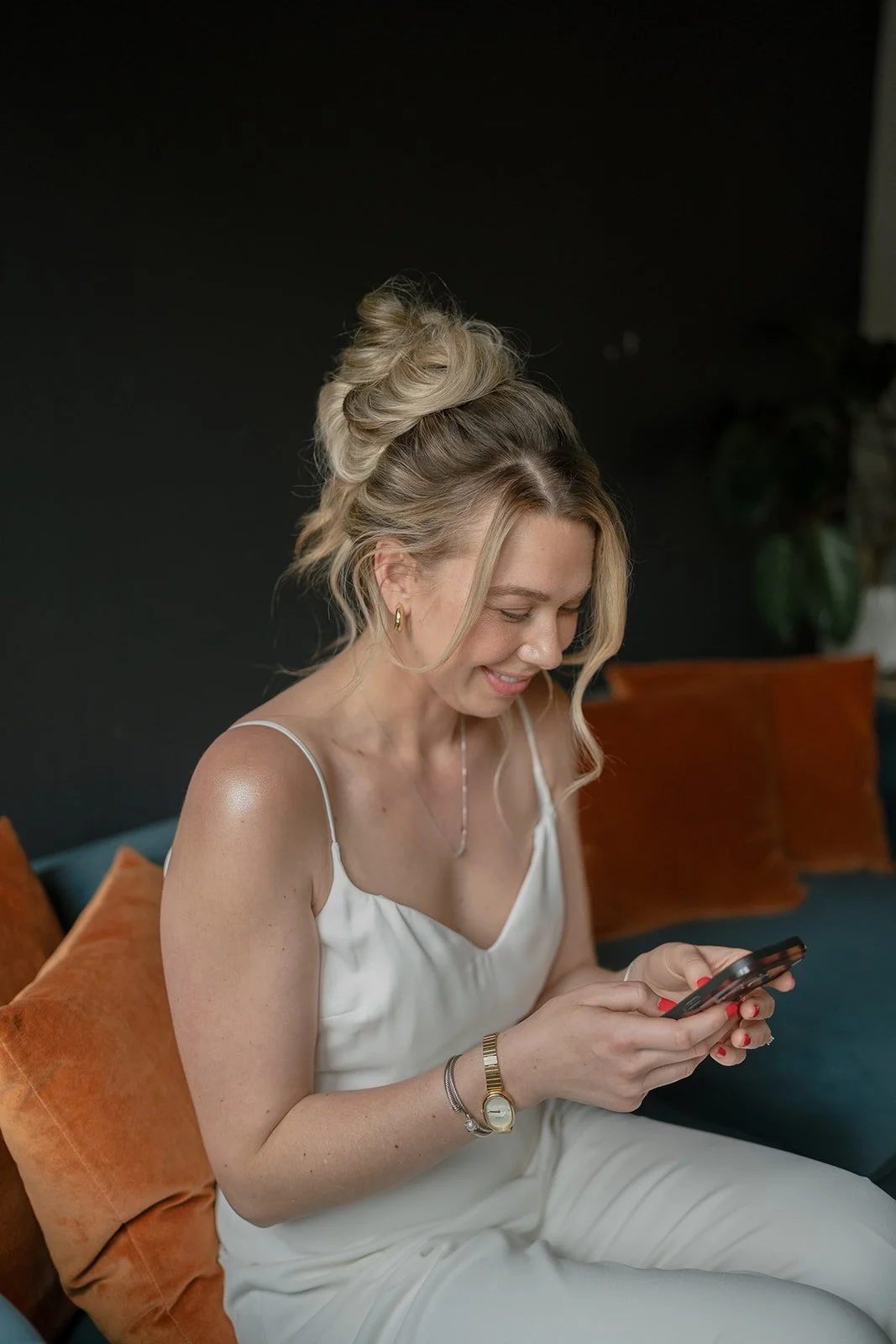 A woman wearing a white dress, sitting on a couch with orange pillows, smiling while looking at her phone, with a black wall and green plant in the background.