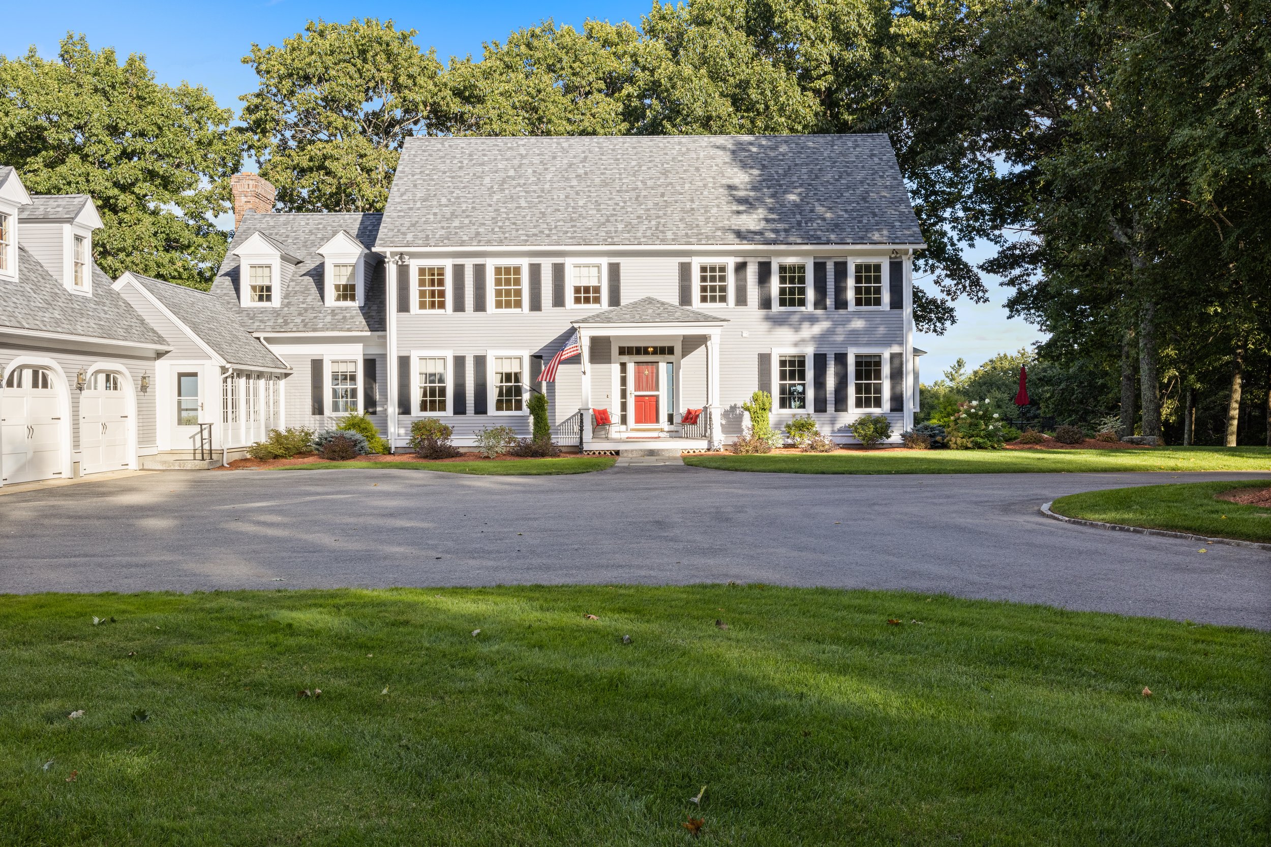 Large colonial-style house with gray siding, black shutters, and a red front door, surrounded by green lawn and trees, with a curved driveway leading to the main entrance.