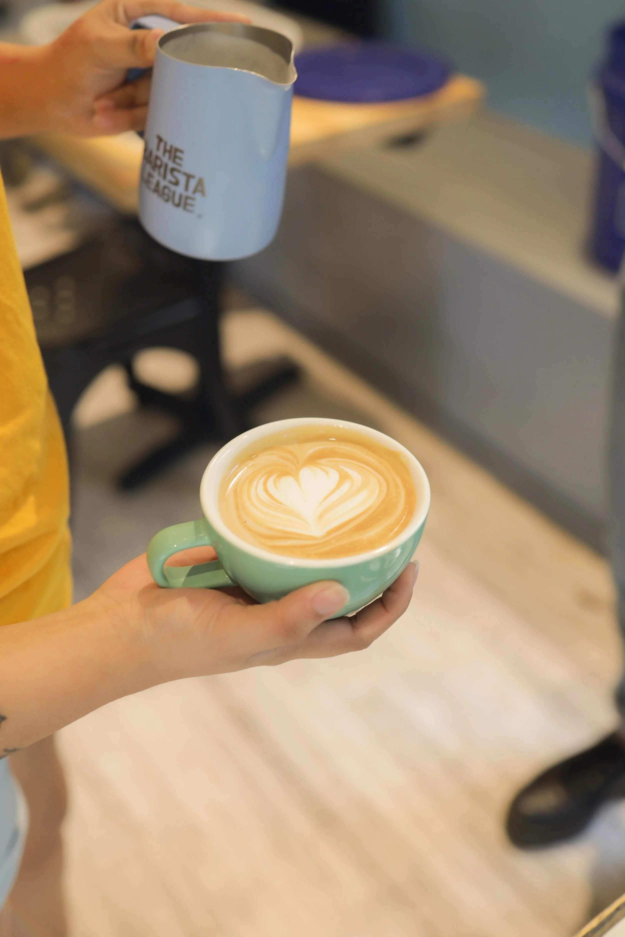 A person holding a bamboo mug that says 'The AISTA League' in one hand and a green ceramic cup with latte art in the other.