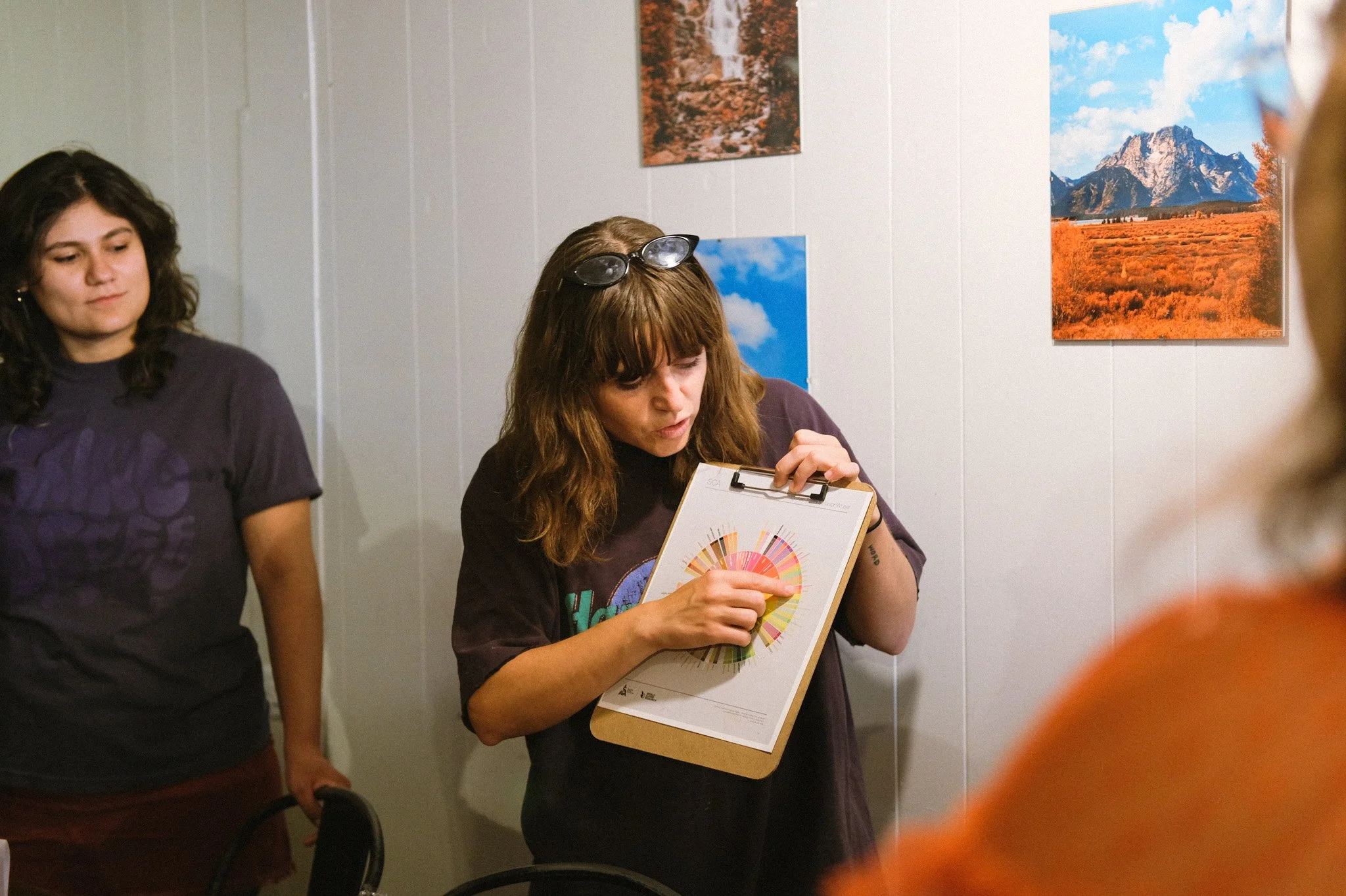A woman with brown hair and sunglasses on her head holds a clipboard with a colorful chart, explaining or presenting it to a group. Two other women are listening, with one on the left wearing a dark shirt and another partially visible on the right. There are landscape photographs on the white wall behind them.