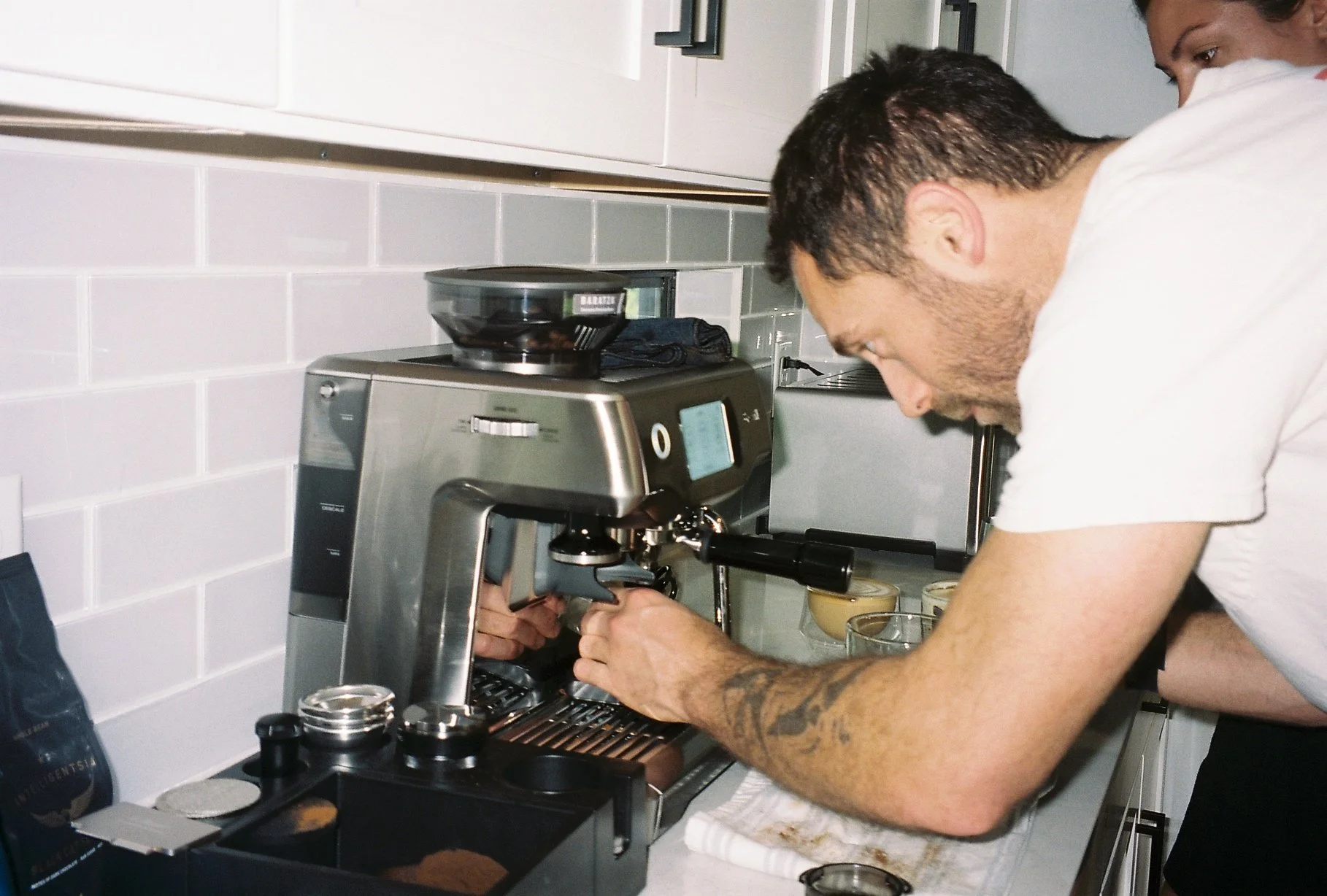A man making coffee with an espresso machine in a kitchen while two women observe behind him.