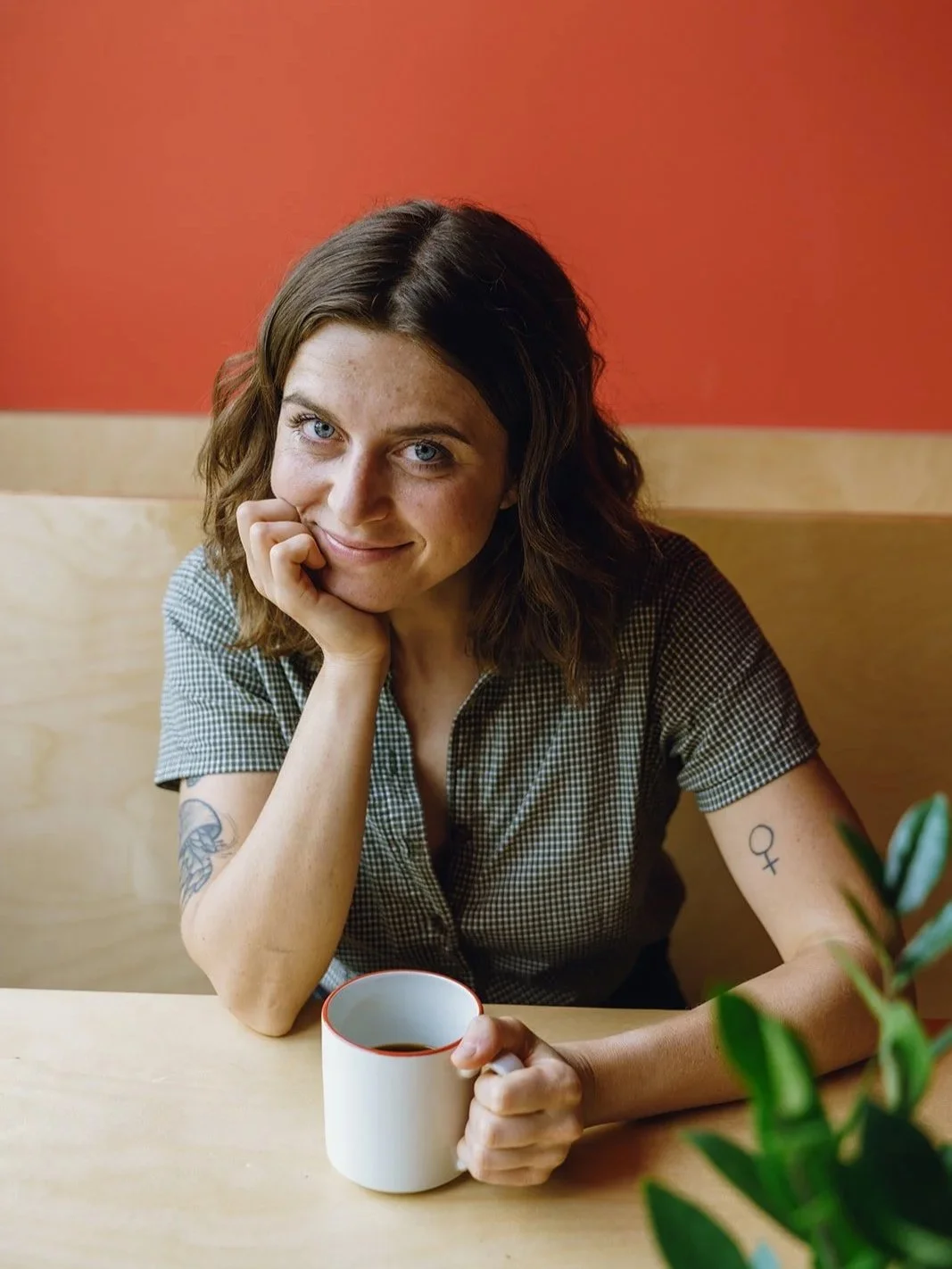 A woman with wavy brown hair and light blue eyes sitting at a wooden table, holding a white mug with a red rim, smiling softly, with a potted plant partially visible in the foreground, and a red wall in the background.
