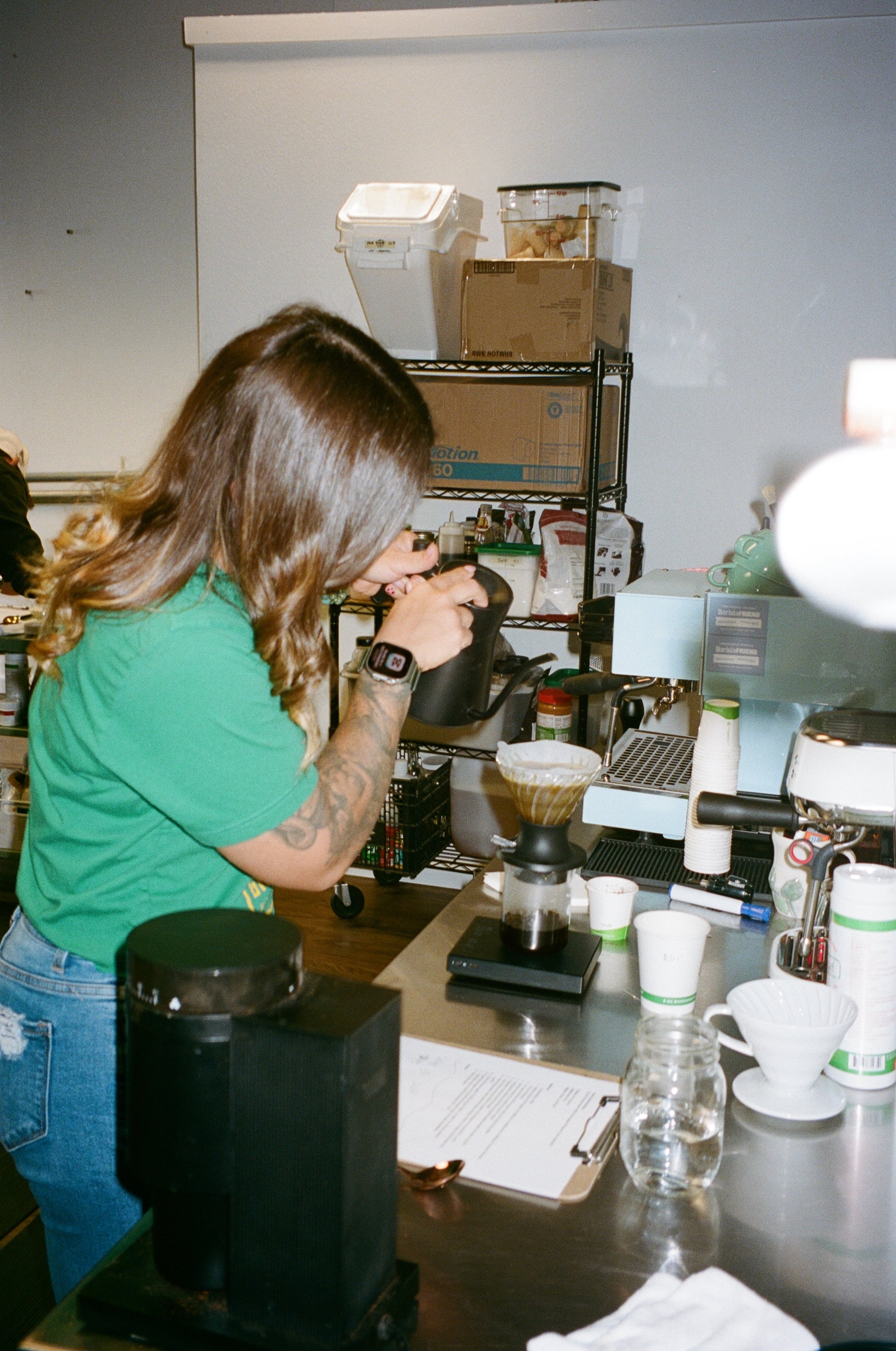 A woman with long brown hair, wearing a green shirt, making coffee in a coffee shop kitchen, surrounded by coffee equipment and supplies.
