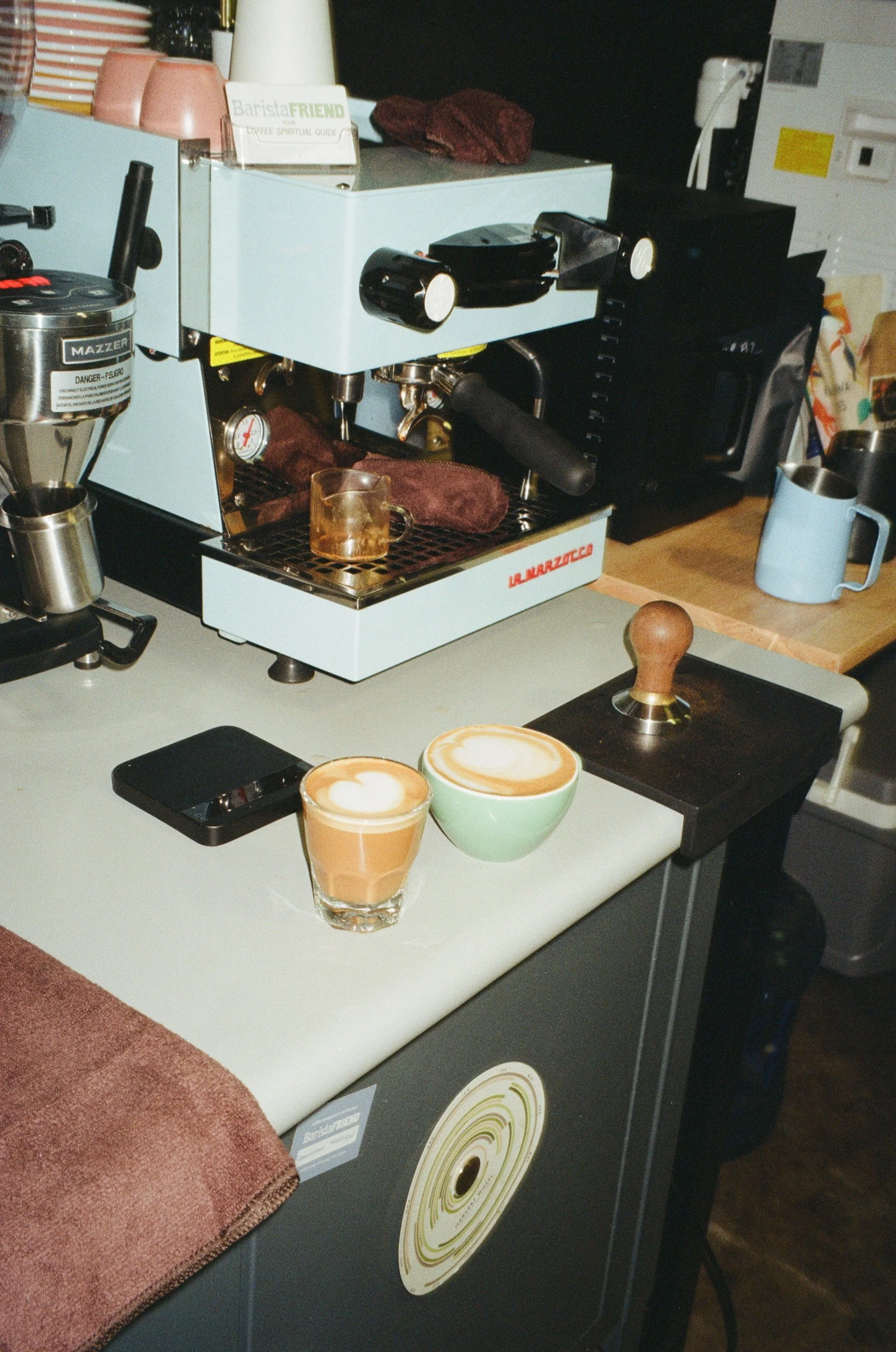 Two cups of coffee with latte art on a white countertop, a coffee tamper, and a scale, with a coffee machine and other kitchen items in the background.