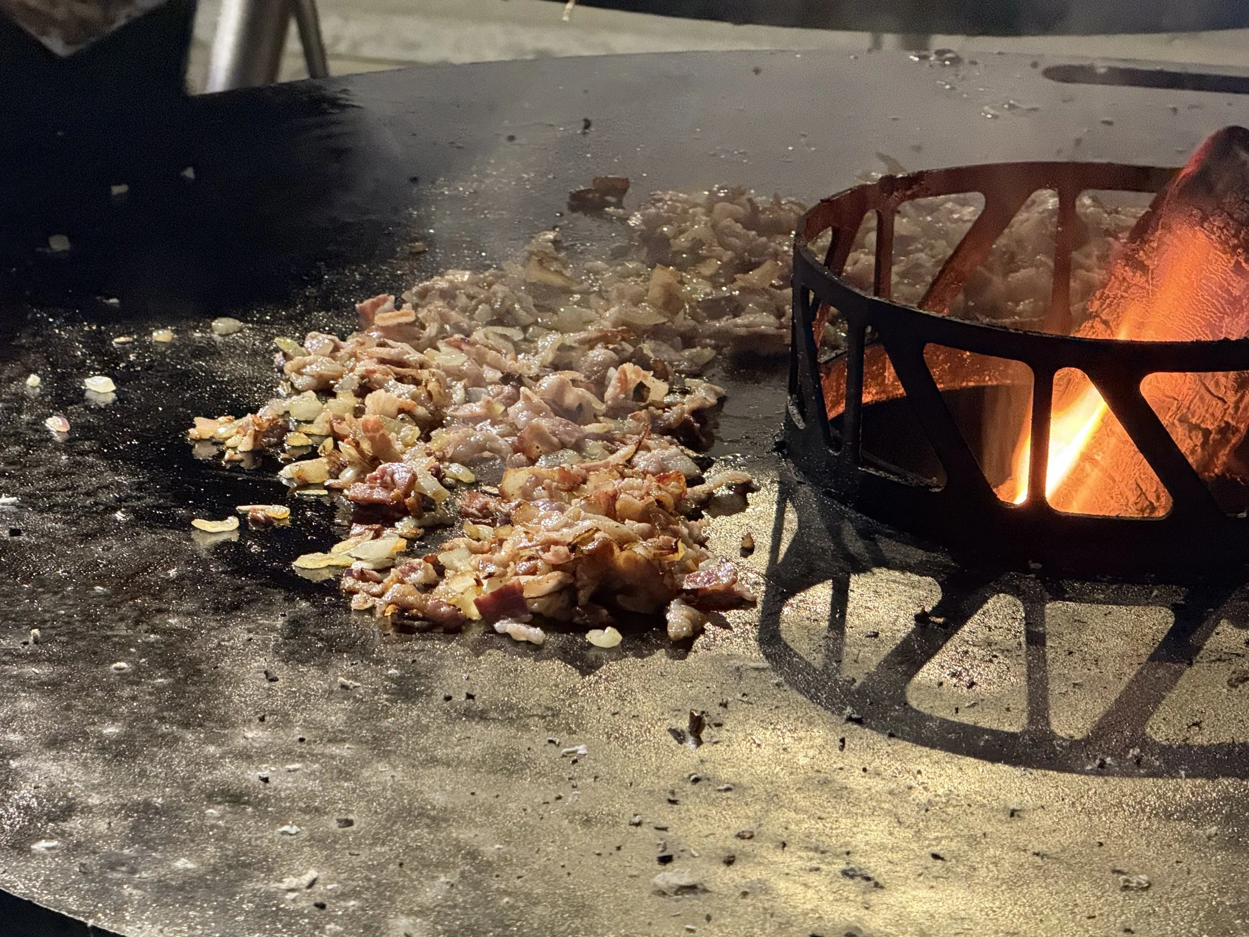 Gebackenes Fleisch und Zwiebeln auf einer heißen Grillplatte, neben einem offenen Feuerbrander.