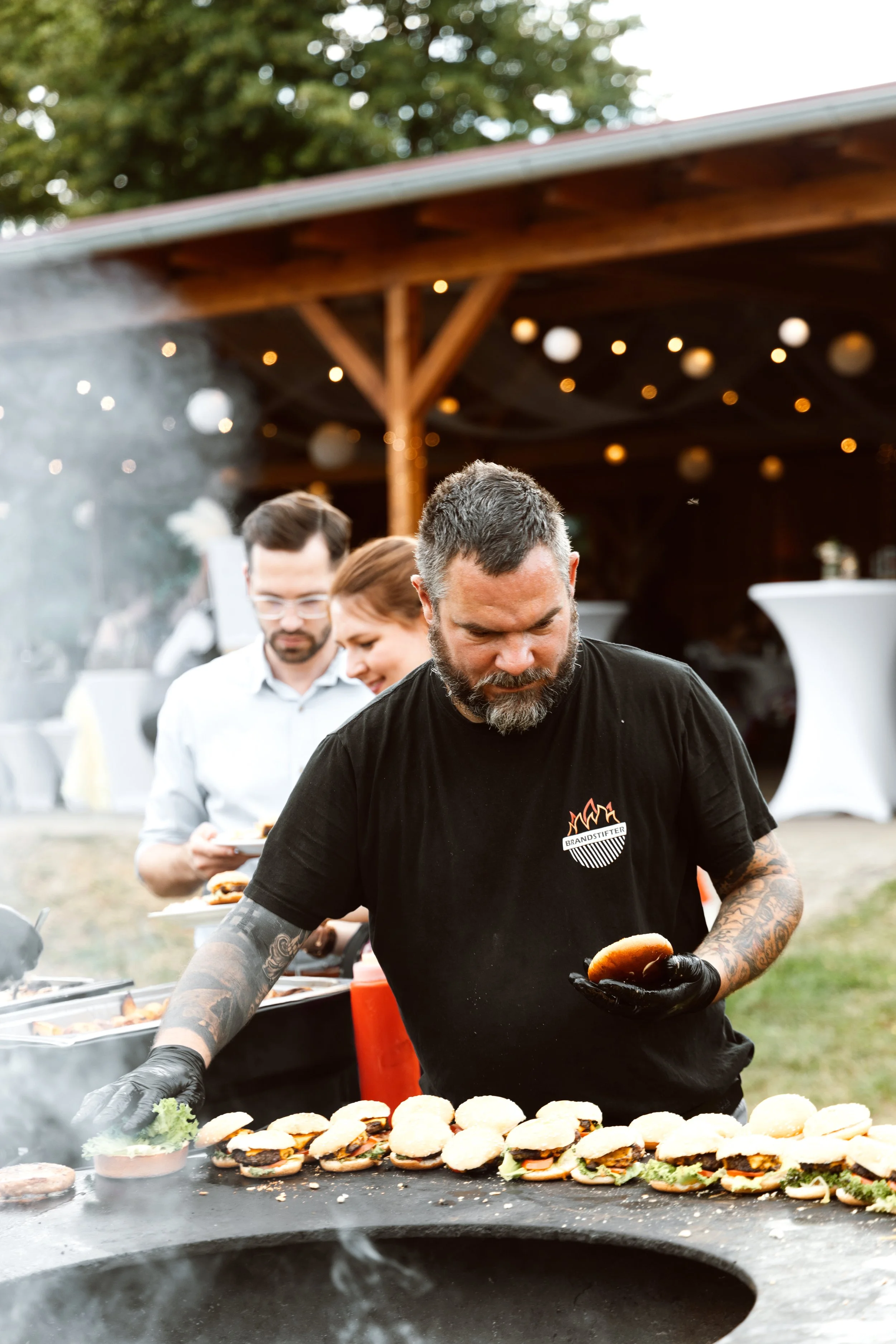Ein Mann macht Sandwiches auf einem Grill bei einer Feier im Freien, im Hintergrund stehen Gäste in einer Umgebung mit Lichterketten.