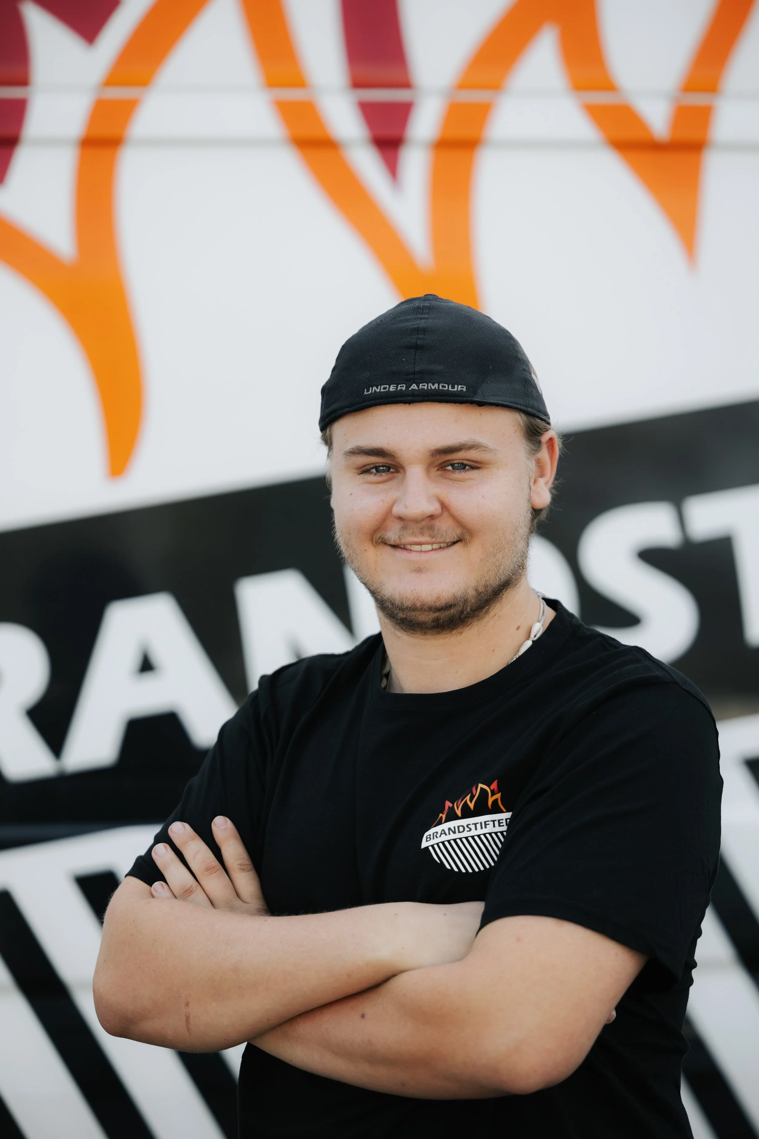 Ein junger Mann steht vor einem Whiteboard mit Feuerwehr-Logo, trägt ein schwarzes T-Shirt mit Brandschutz-Logo und eine schwarze Kappe, lächelt und hat die Arme verschränkt.