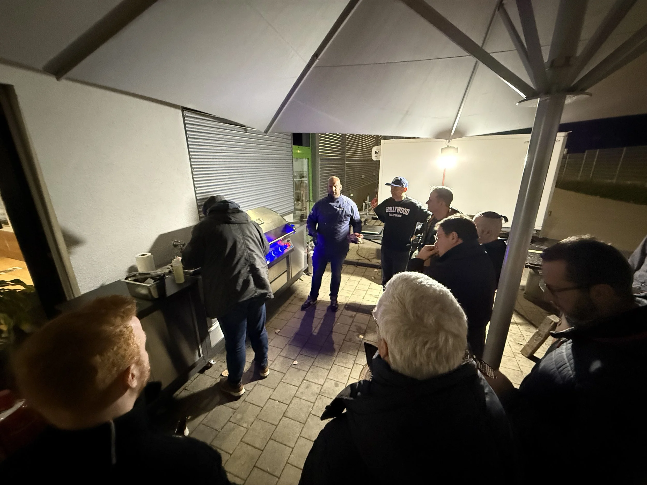 Gruppe von Menschen beim Essen im Freien an einem Abend, unter einem Sonnenschirm, mit einem Grill und einem Whiteboard im Hintergrund.
