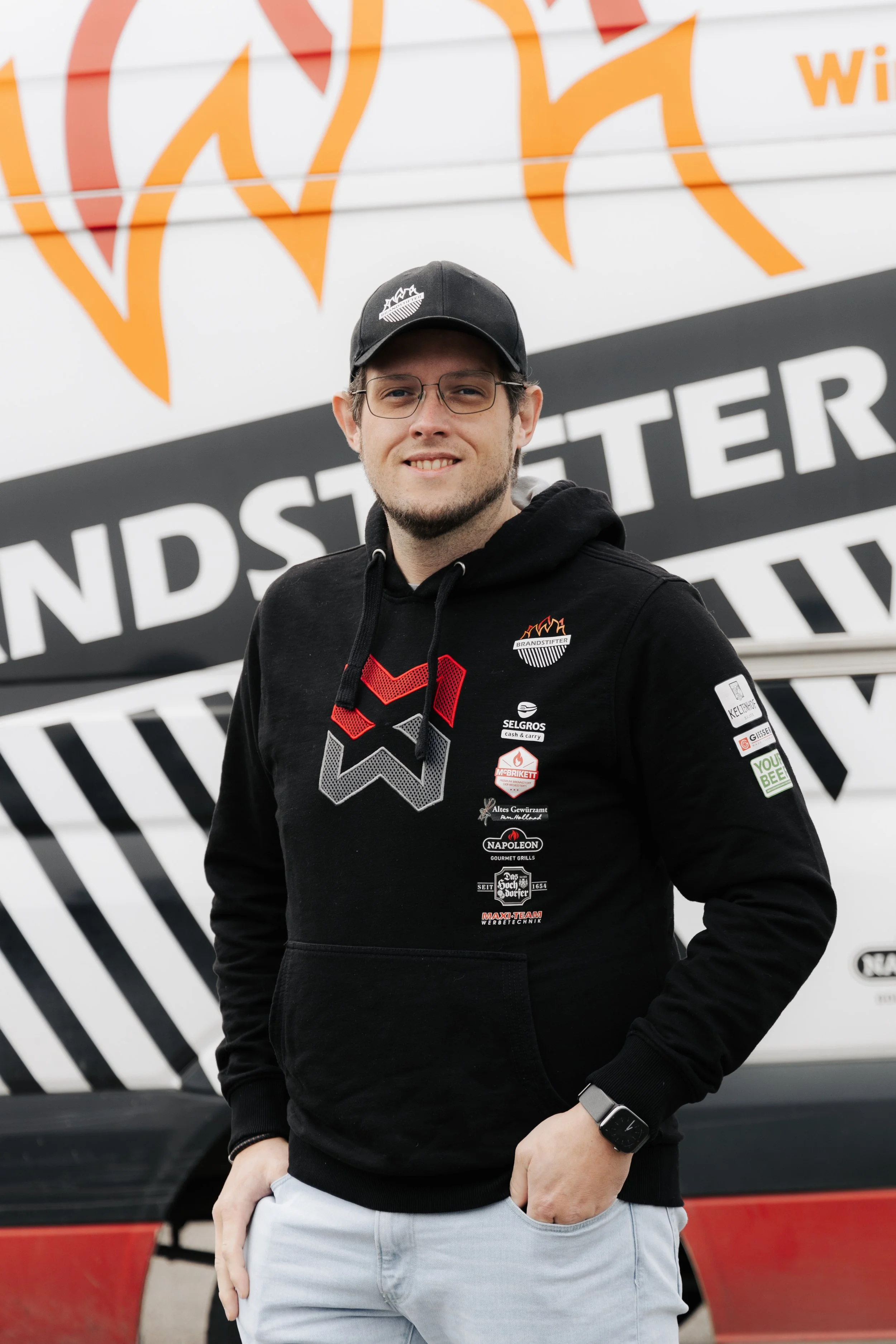 Junger Mann trägt schwarze Kapuzenjacke mit Logos, schwarzen Hut, Brille und Smartwatch, steht vor einem Fahrzeug mit großen Druckaufklebern, die Austragungsort "Landstede" und Logo "Branderst" zeigen.