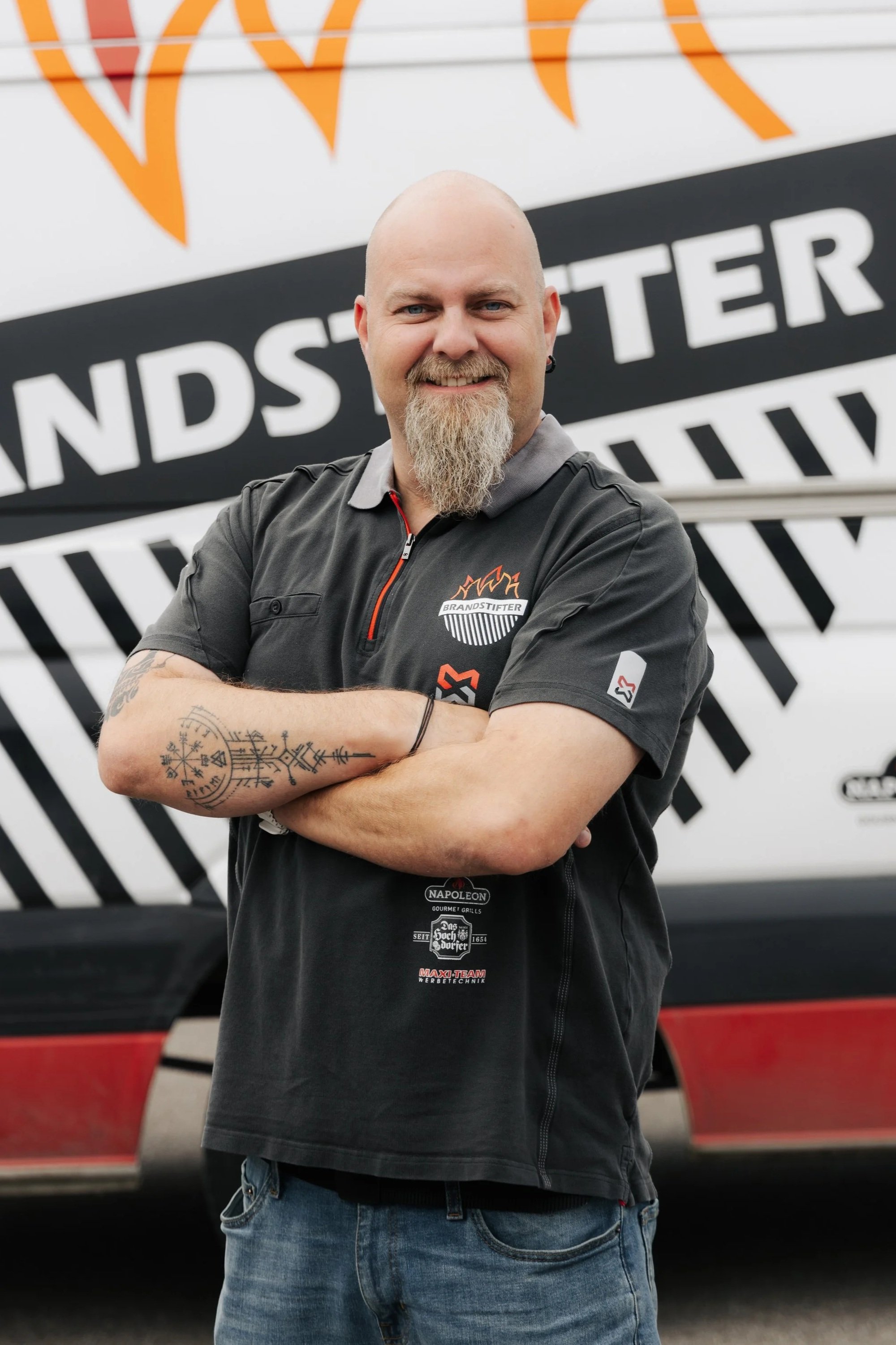 Ein Mann mit einem grauen Bart, tätowiertem Arm, trägt ein schwarzes T-Shirt mit Logos und steht vor einem Fahrzeug mit einem großen Logo, das 'BRANDSTIFTER' enthält.