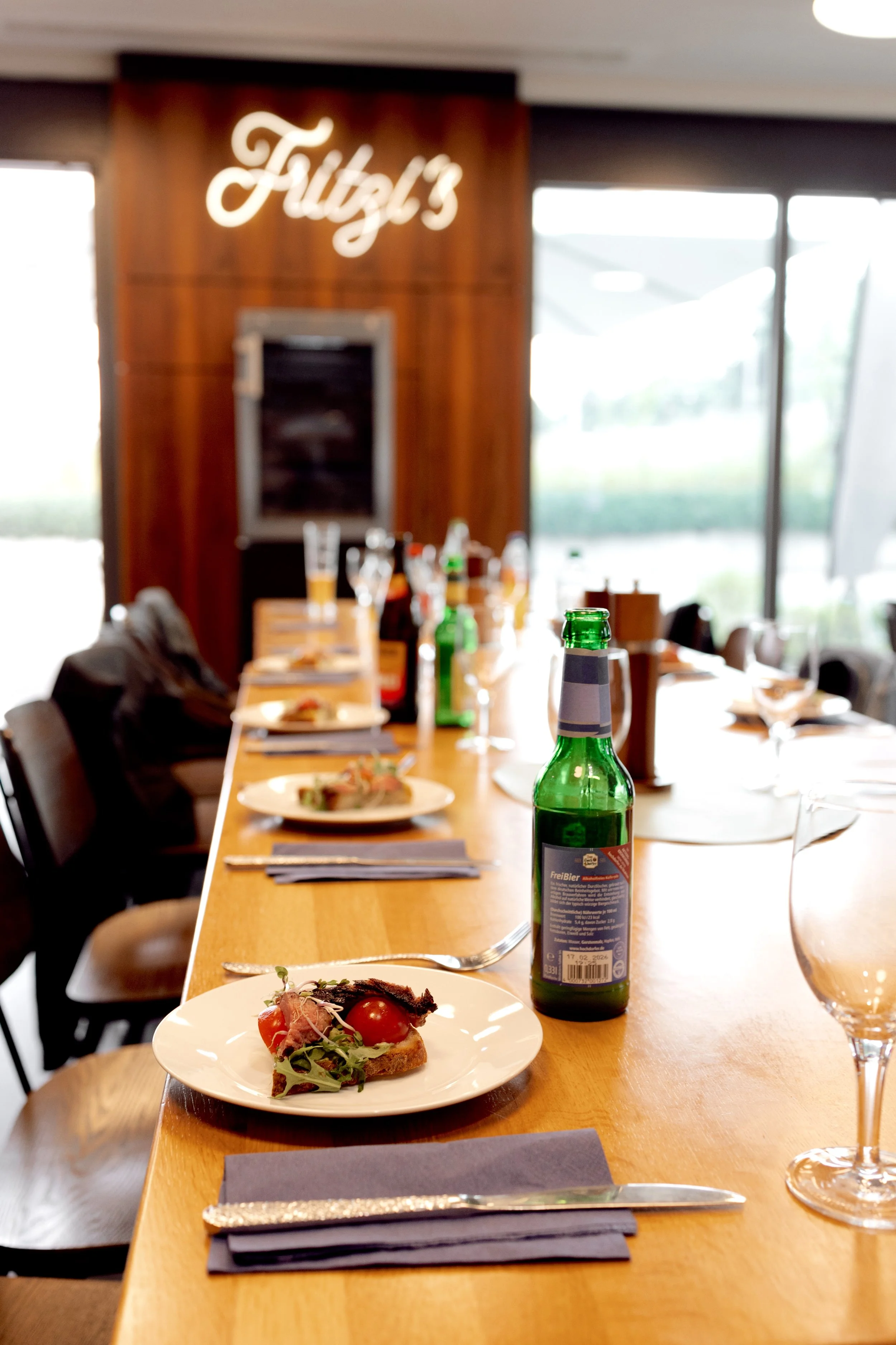 Ein Restaurant mit einem langen Holztisch, belegten Tellern und Getränken. Im Hintergrund eine Wand mit der Aufschrift 'Fitz's' und große Fenster, die Tageslicht hereinlassen.