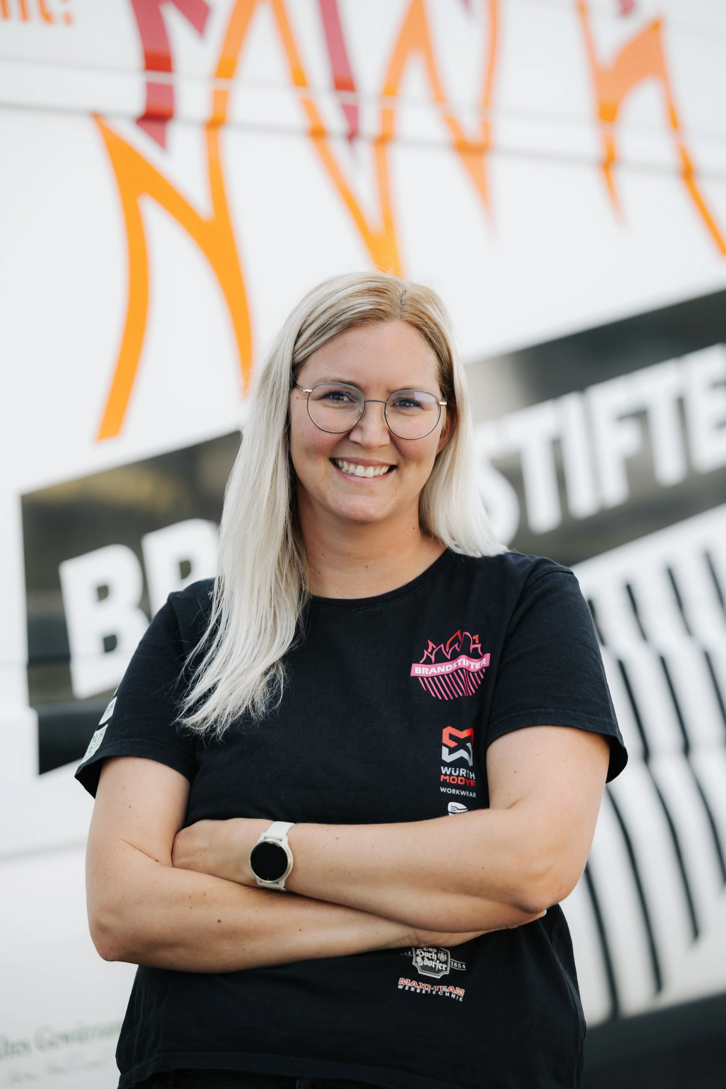 Eine lachende Frau mit blonden Haaren und Brille trägt ein schwarzes T-Shirt mit Logos und steht vor einem großen Banner mit orangefarbenen und weißen Schriftzügen.