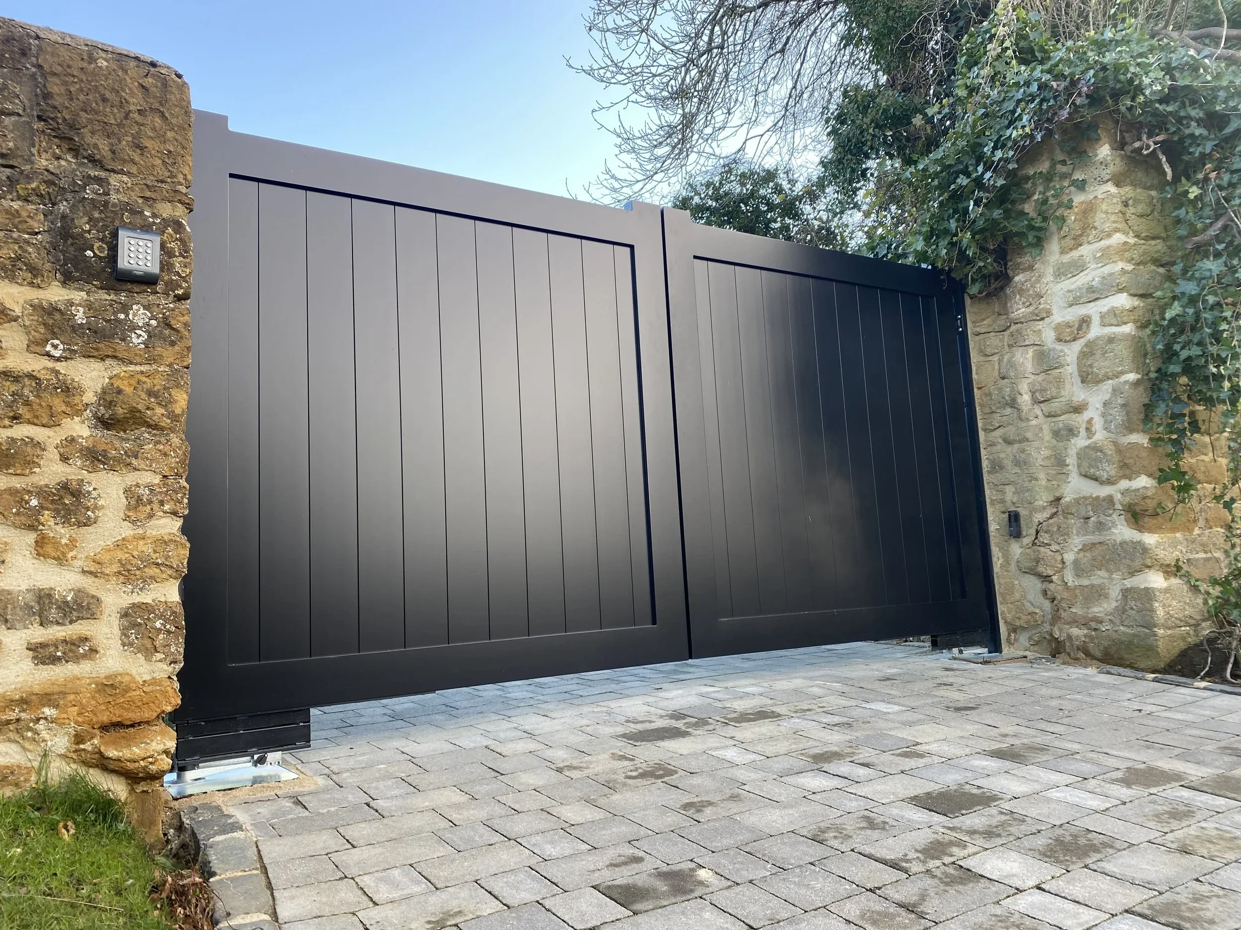 Black modern gate installed between stone walls with metal keypad on the left stone wall, paving stones on the ground, surrounded by greenery and bushes, under a clear sky.