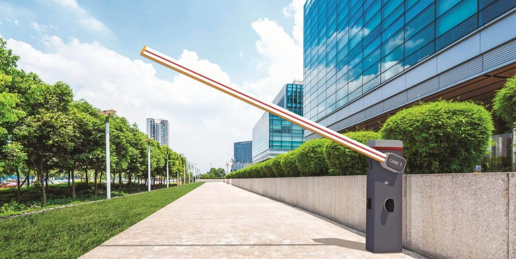Modern city sidewalk lined with green trees and shrubs, with a glass office building on the right and high-rise buildings in the background. A parking garage barrier gate is in the foreground.