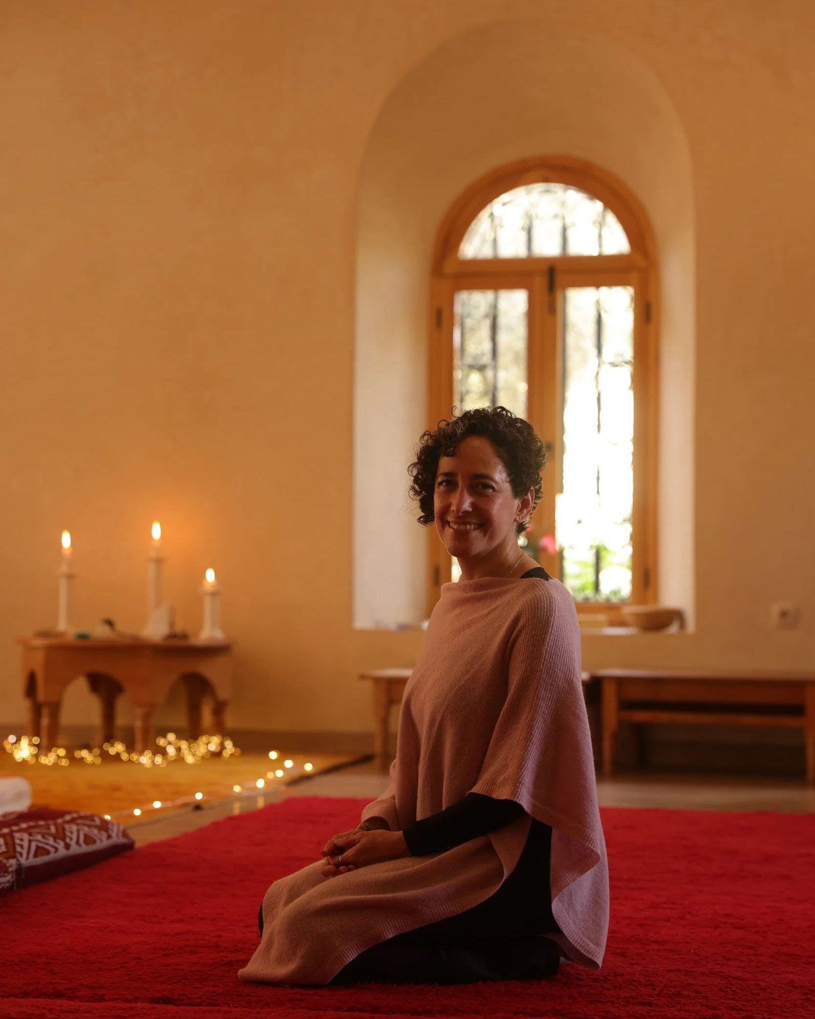Une femme assise en tailleur sur un tapis rouge dans une pièce aux murs beige, avec une fenêtre grande et arrondie derrière elle, éclairée par la lumière naturelle. La pièce est décorée avec des bougies allumées et des guirlandes lumineuses.
