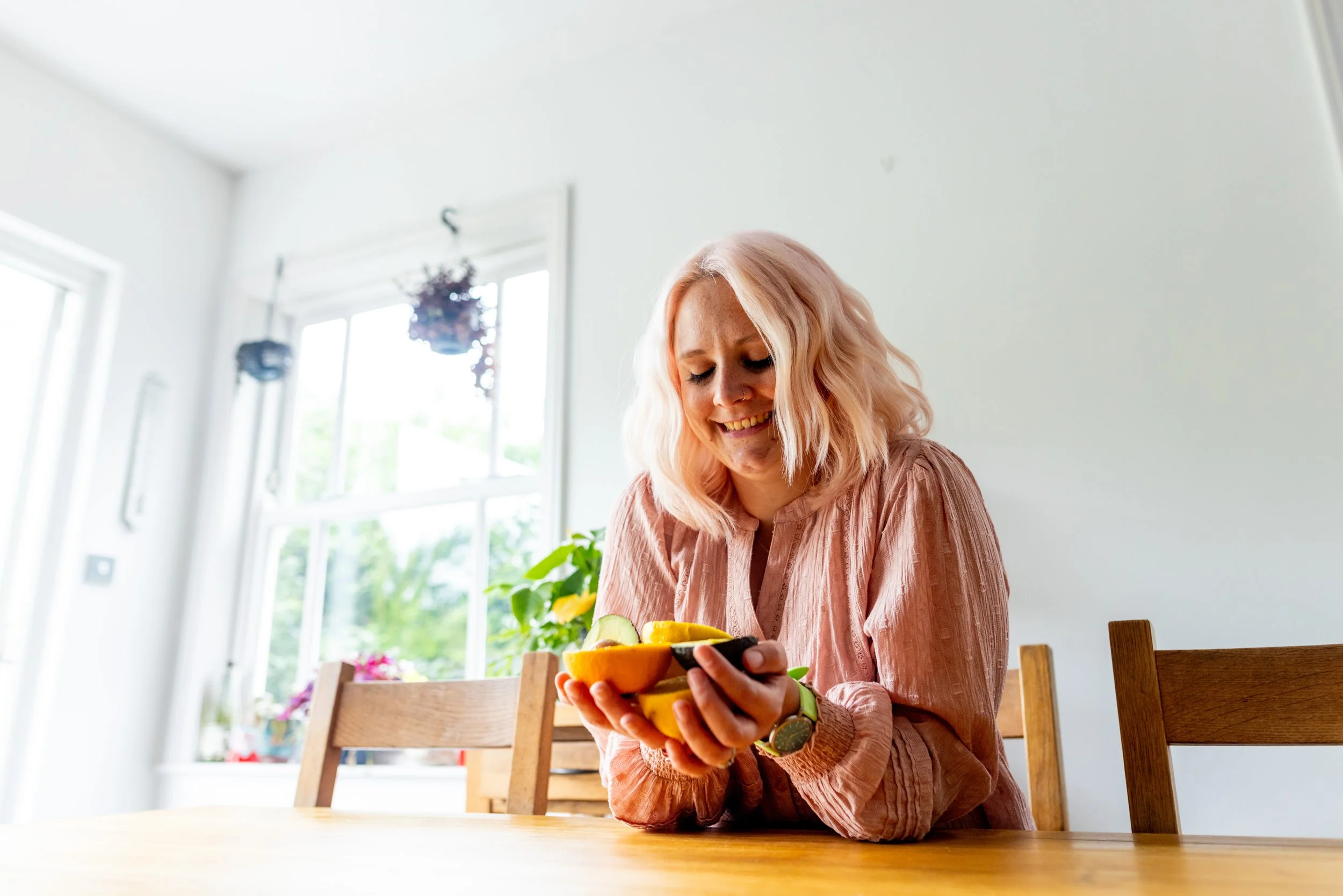 Ruth is sat at a wooden dining table, wearing a pink blouse, holding cut oranges, lemons and limes and looking down at them, smiling
