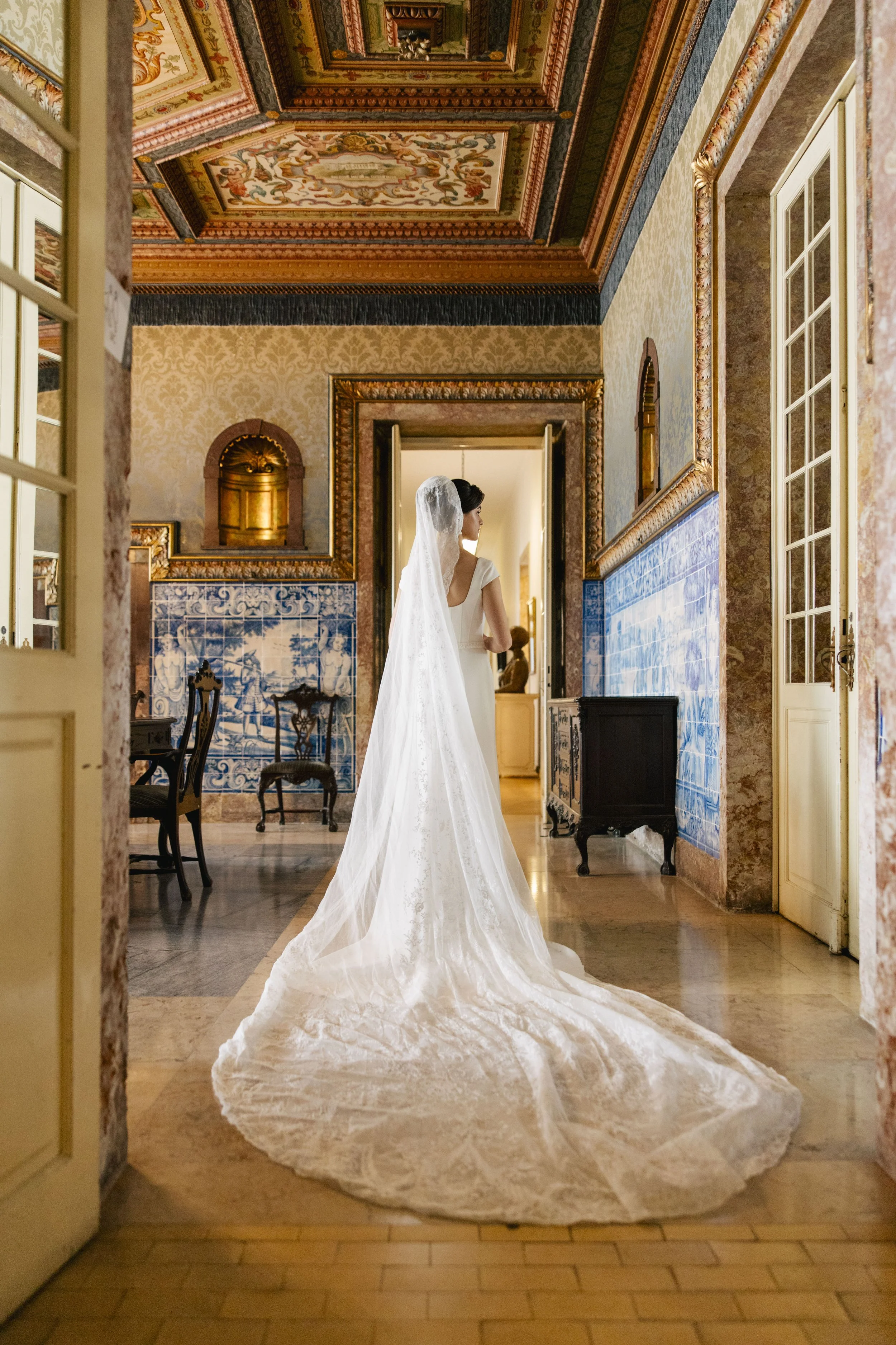 A bride in a white wedding dress with a long train and veil standing in an ornately decorated room, facing away from the camera, with an open doorway behind her and patterned wallpaper on the walls.