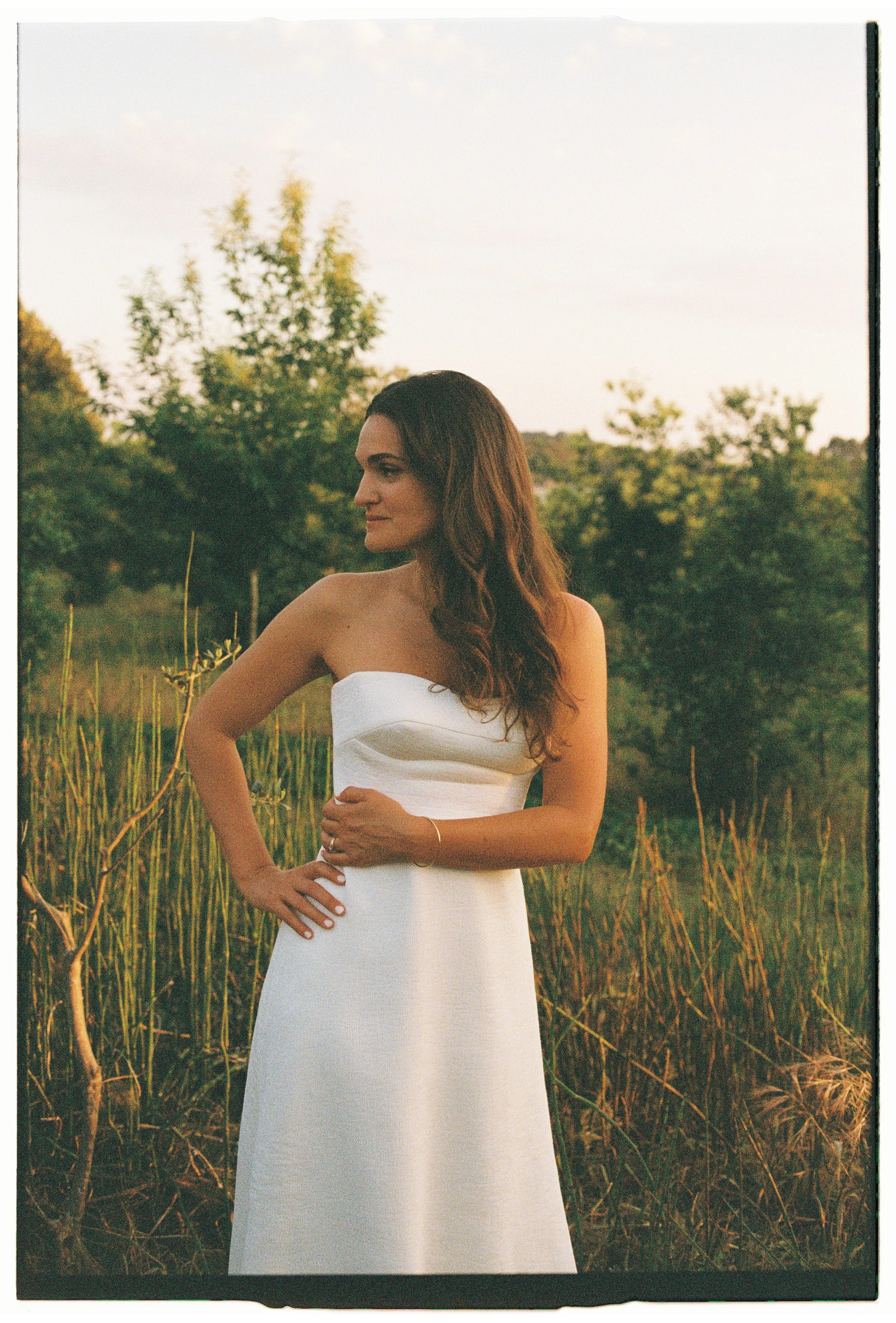A woman with long brown curly hair wearing a strapless white dress standing in a field of tall grass with trees in the background, illuminated by warm golden light.