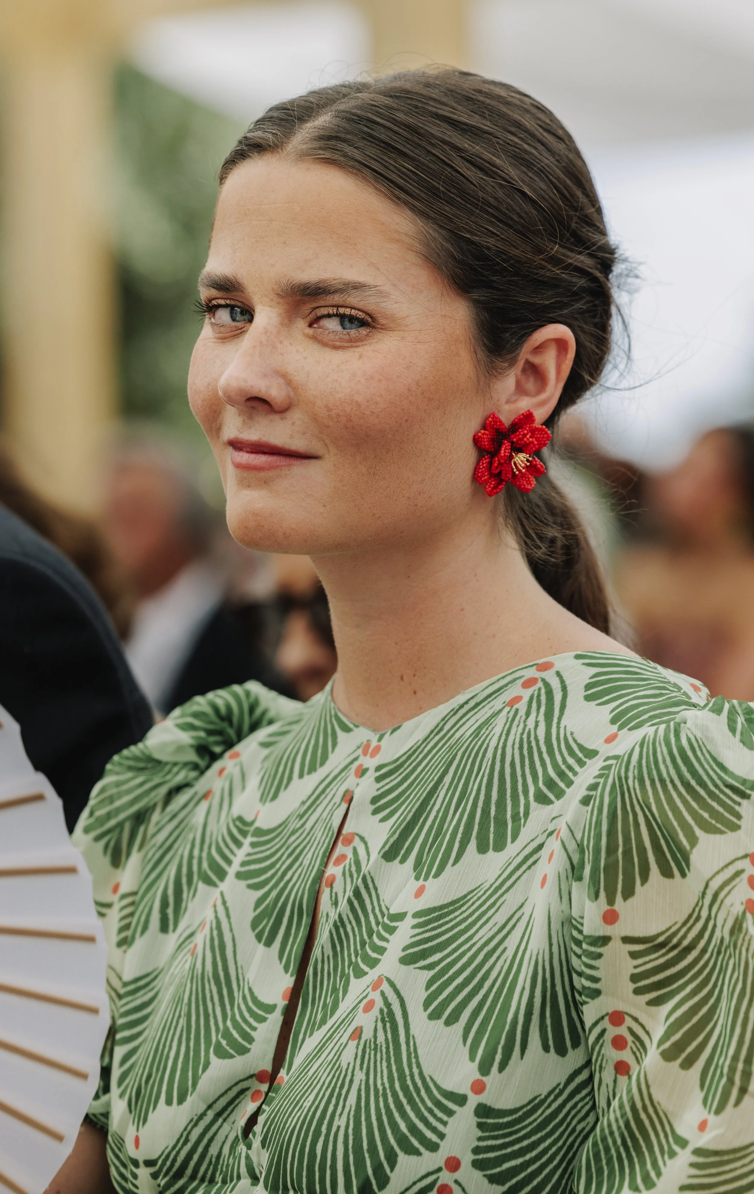 Close-up of a woman with light skin and blue eyes, wearing red floral earrings and a green patterned dress, smiling slightly and looking to her left. Her hair is tied back and she is in a bright, indoor setting with a blurred background.