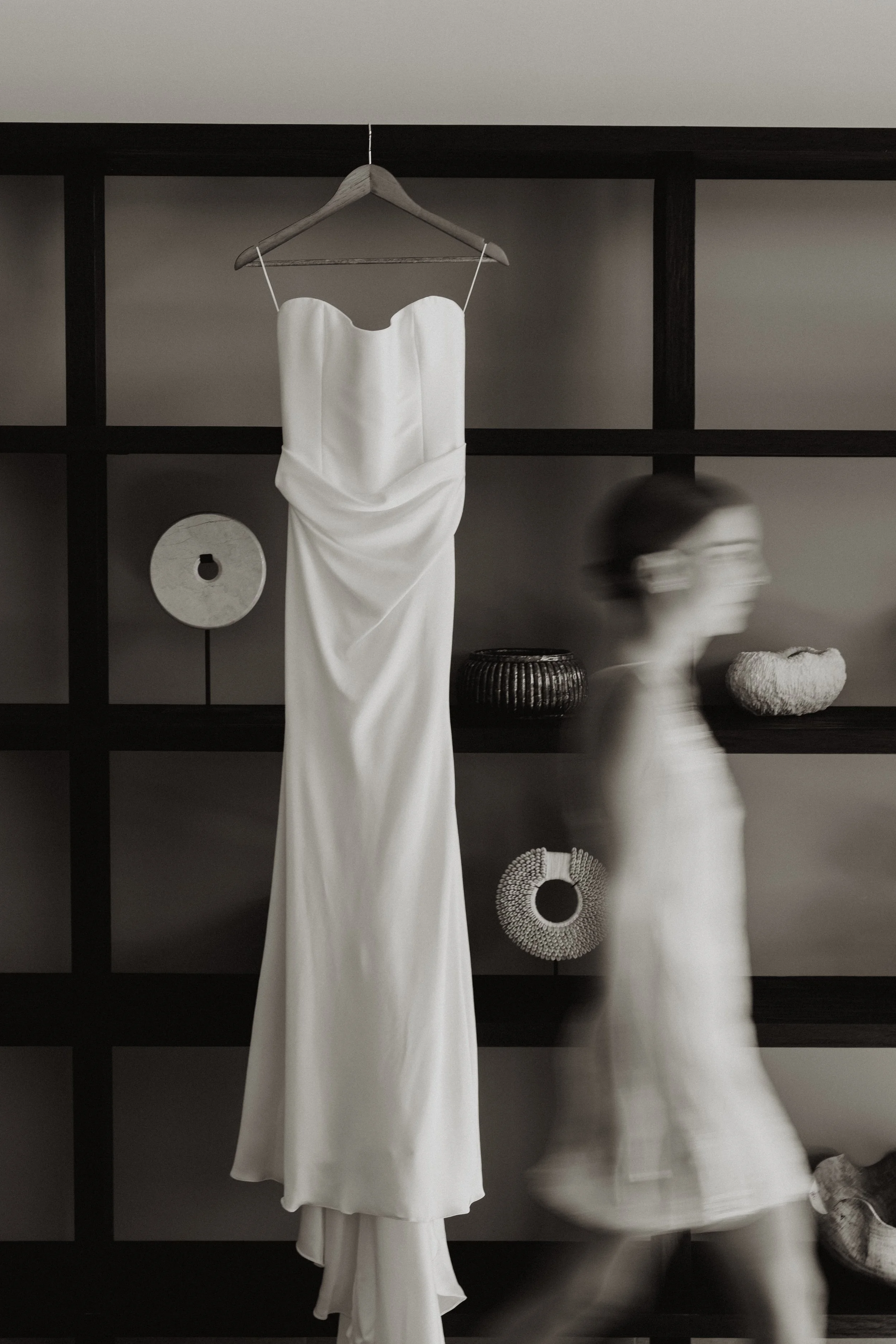 A white wedding dress hanging on a wooden hanger on a black shelving unit, with a blurred person in white walking past it.