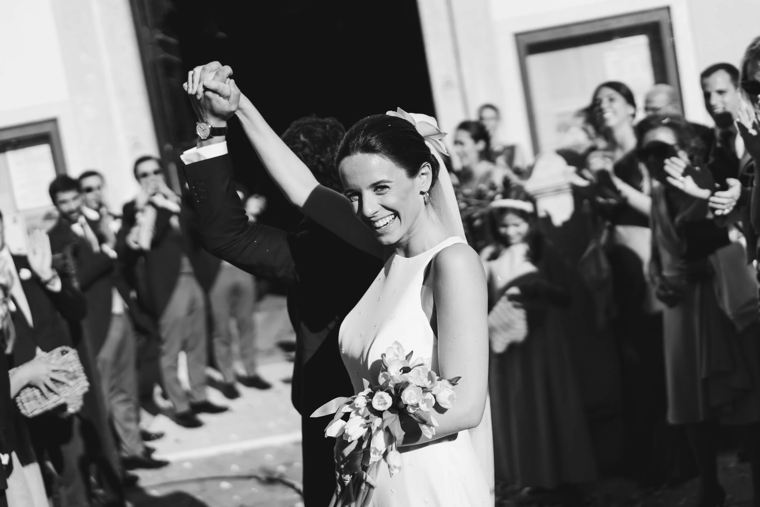 Black and white photo of a bride holding a bouquet of flowers, embracing her partner while dancing with a crowd watching in the background.