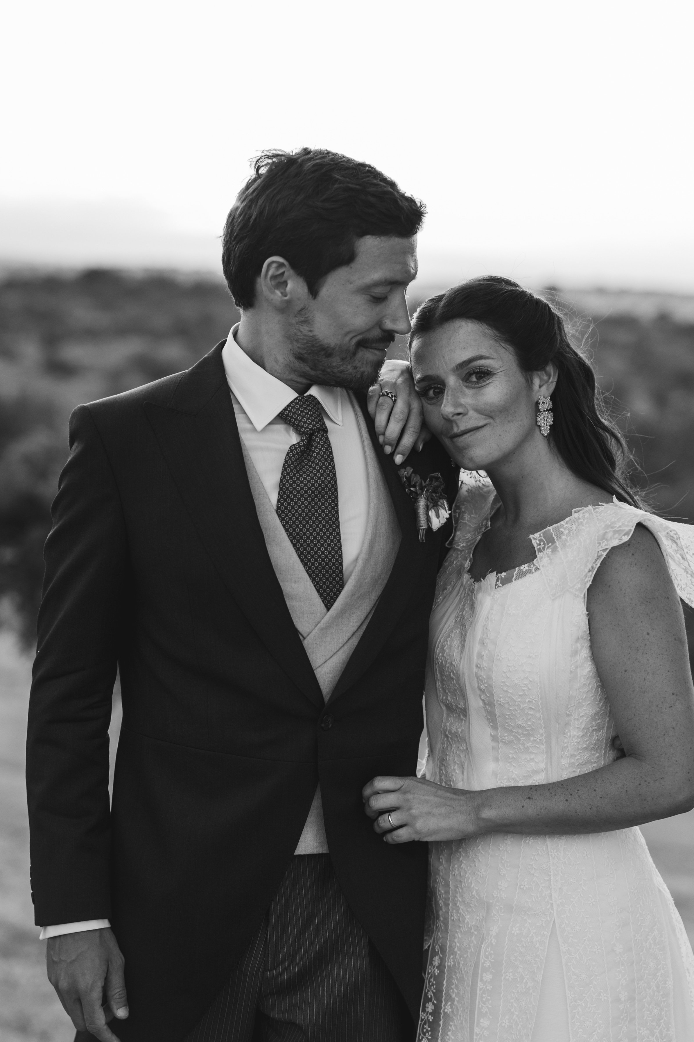 A black-and-white photo of a groom and bride standing close together outdoors, with the groom in a suit and the bride in a wedding dress, both smiling softly and gently touching each other.