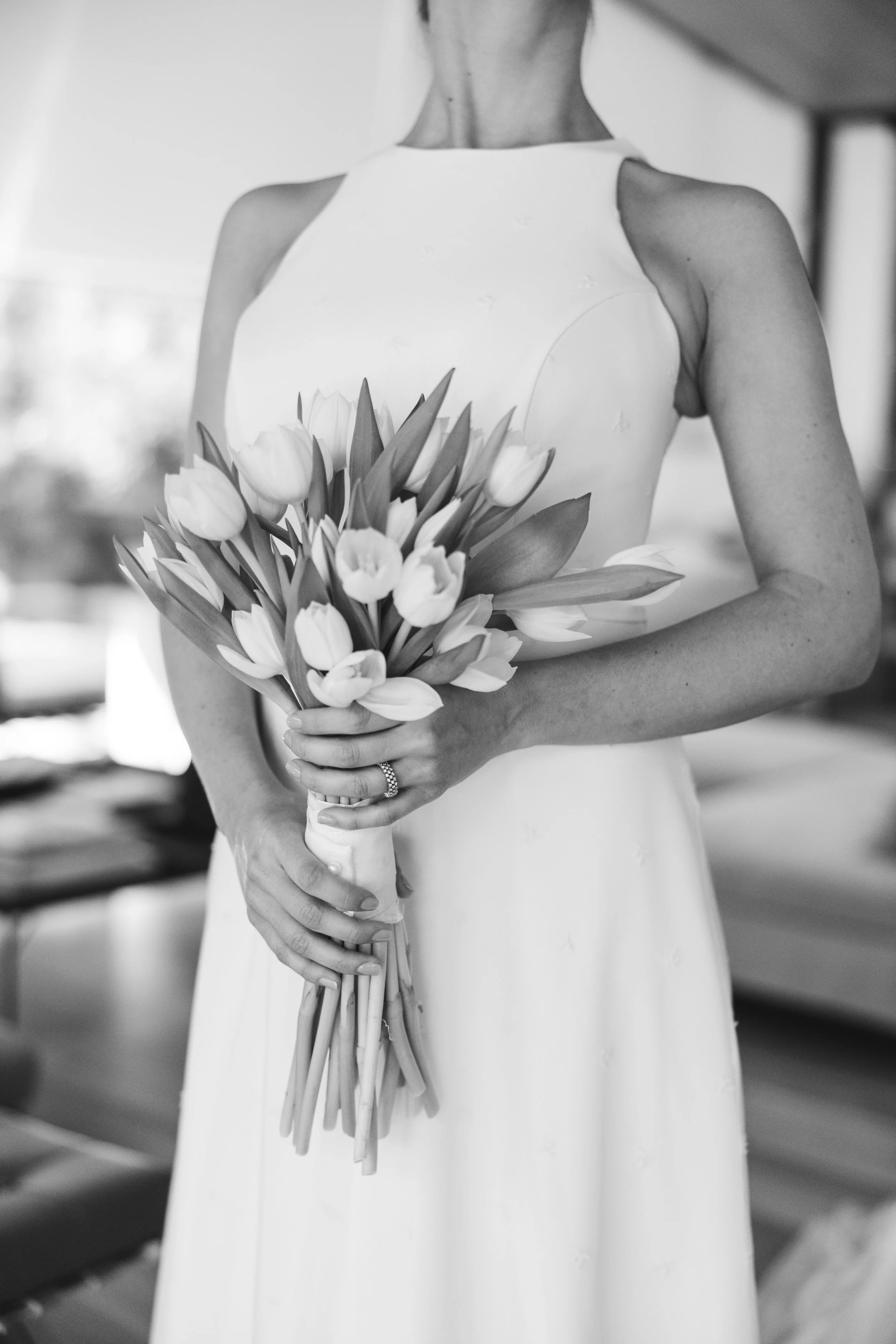 Black and white photo of a woman in a sleeveless dress holding a bouquet of tulips with both hands, wearing a ring, standing indoors.