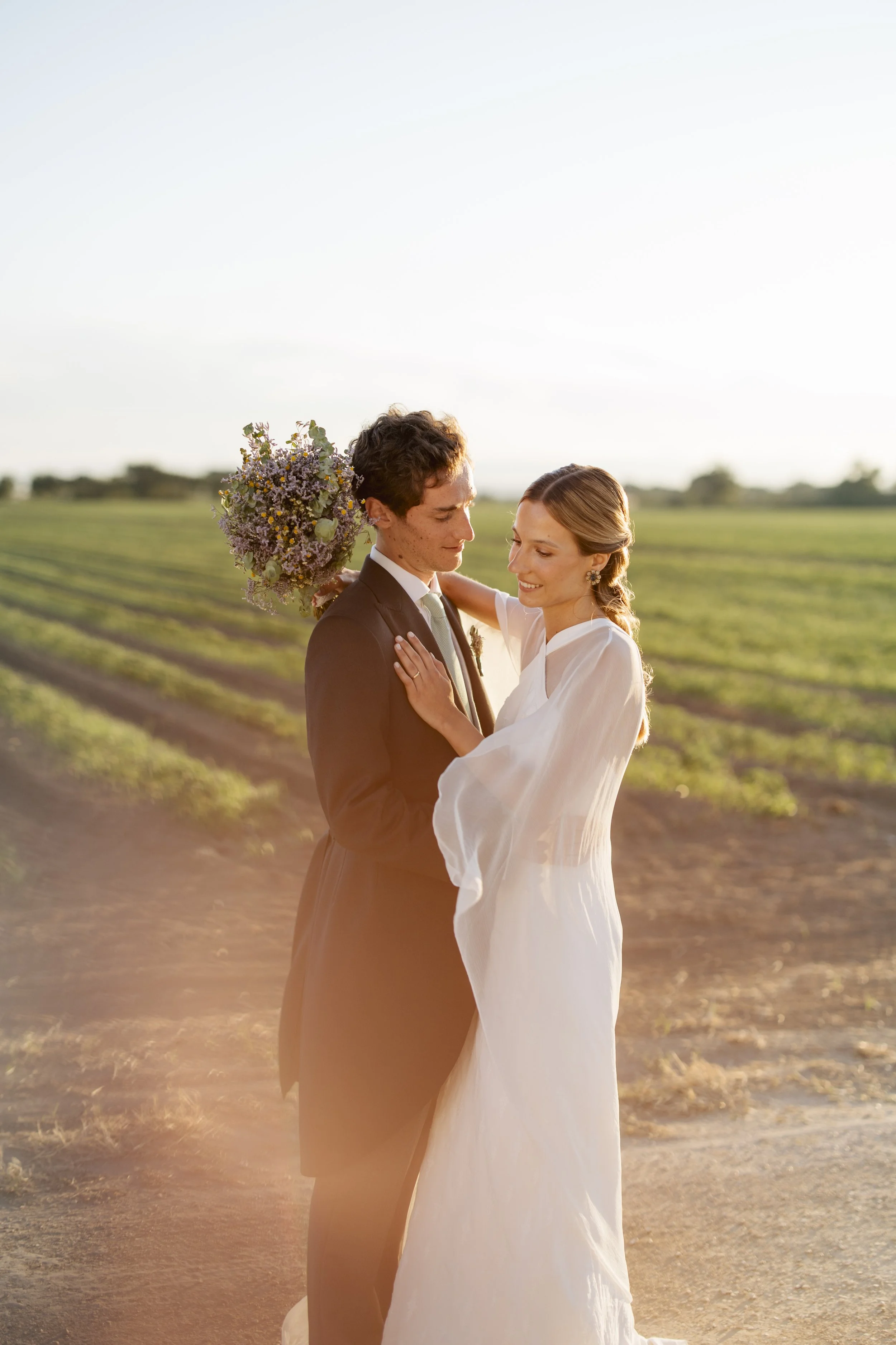 A bride and groom sharing an intimate moment outdoors at sunset, with the bride holding a bouquet and both dressed in wedding attire, standing in a rural field with greenery in the background.
