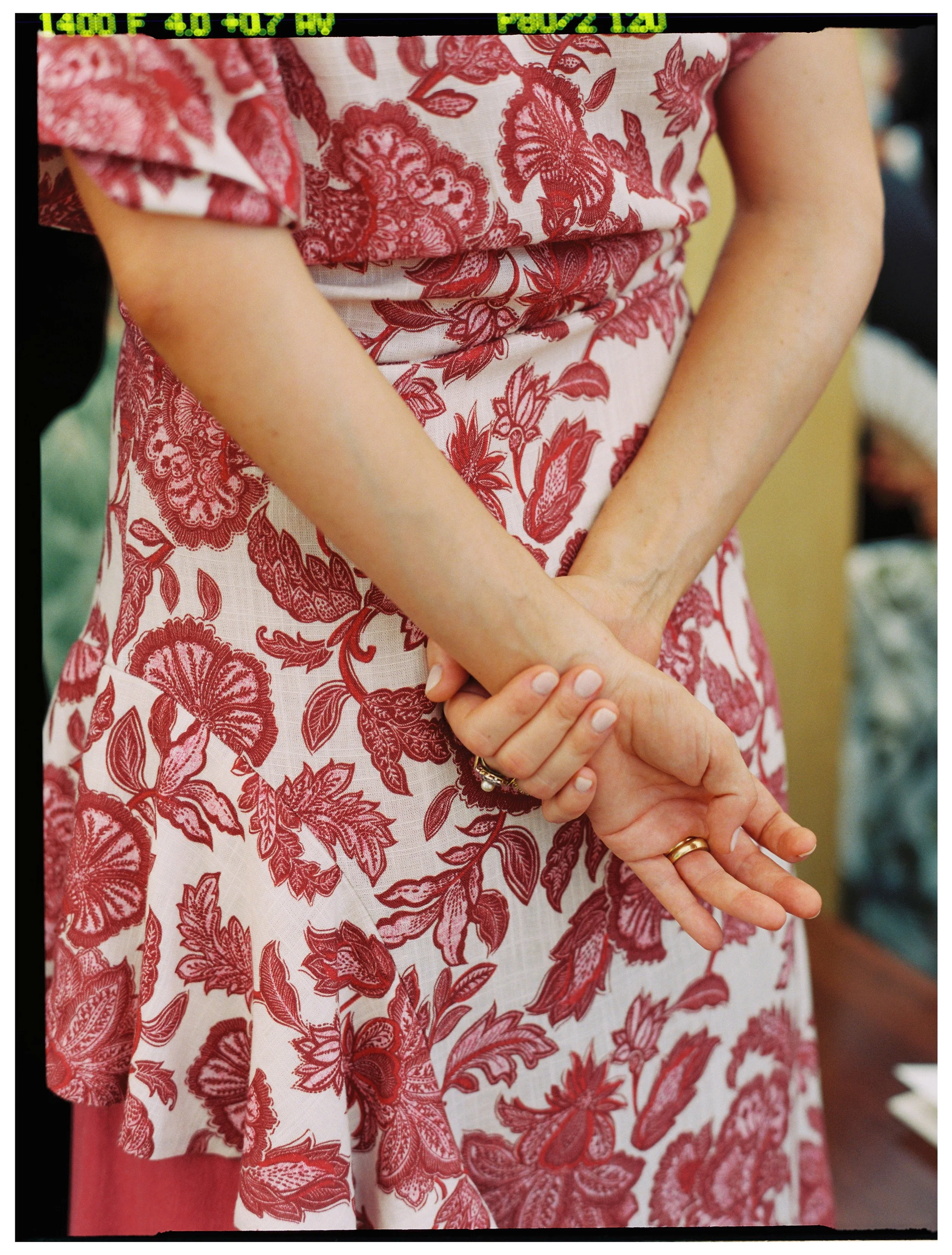 Close-up of a person's hands clasped together, wearing a wedding ring, in front of a red and white floral dress with intricate floral and leaf patterns. The person has light skin and neatly trimmed nails.