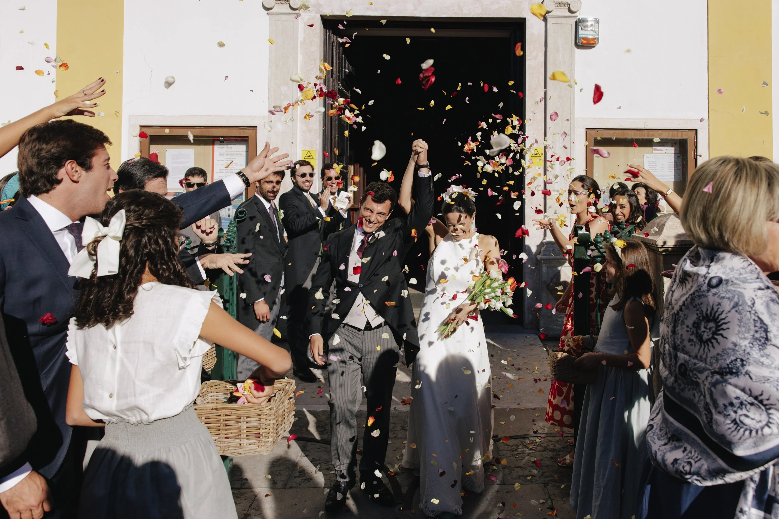 A wedding celebration with the bride and groom exiting a building while guests throw flower petals and celebrate.