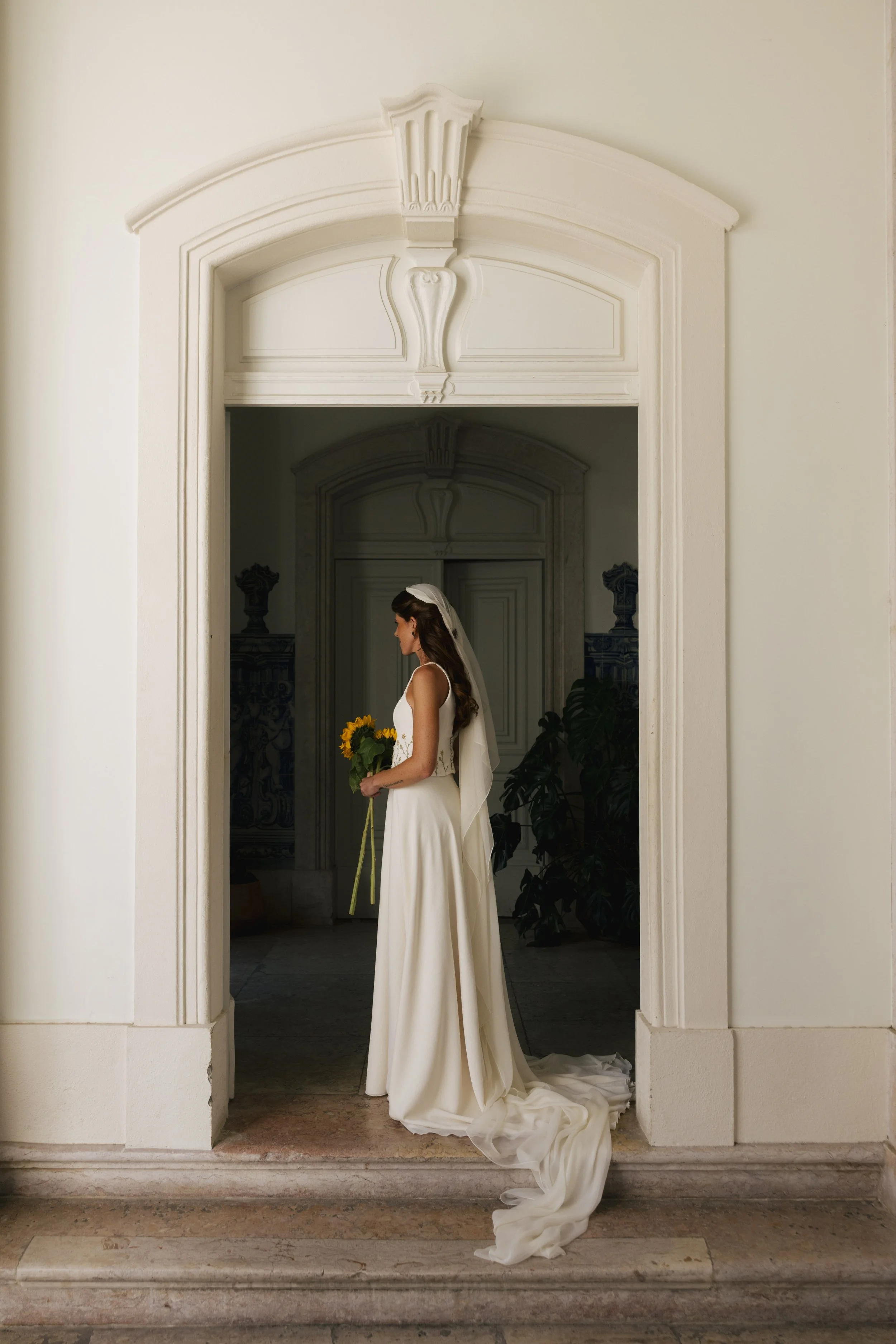 A bride in a white wedding dress holding a bouquet of yellow sunflowers, standing in front of a large ornate white doorway with intricate molding, inside a building with classical architecture.