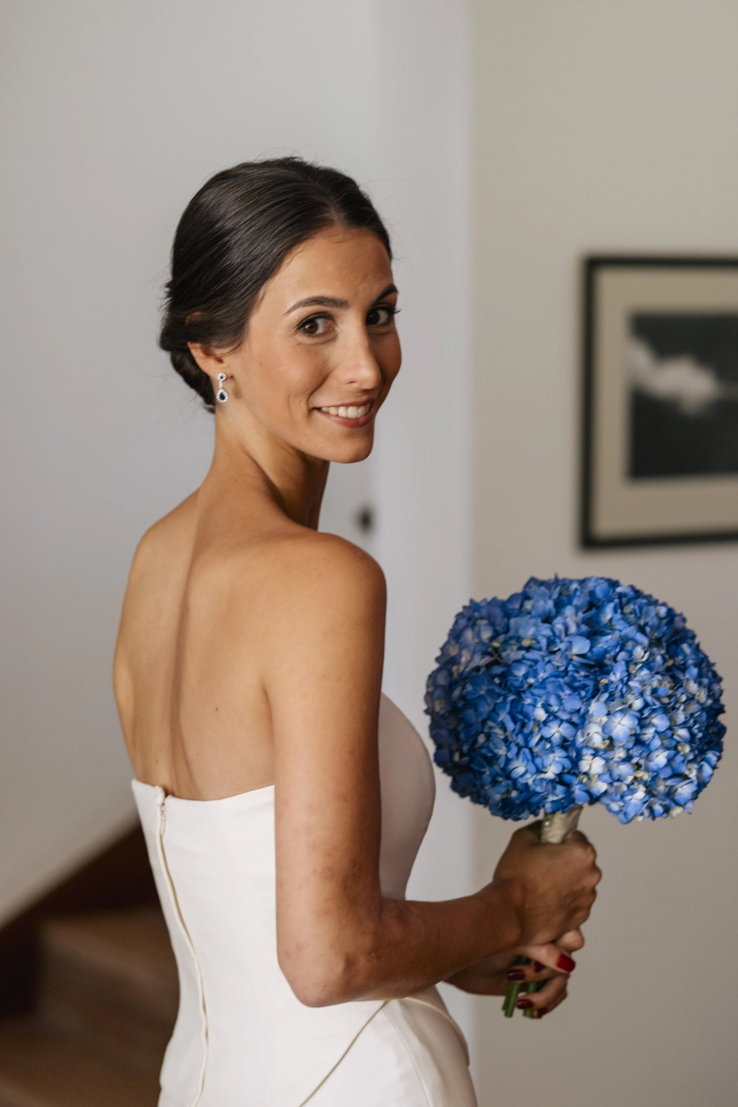 A woman in a strapless white dress holding a bouquet of blue hydrangeas, smiling, with dark hair styled in a sleek updo, and wearing earrings, standing indoors near a framed picture on the wall.