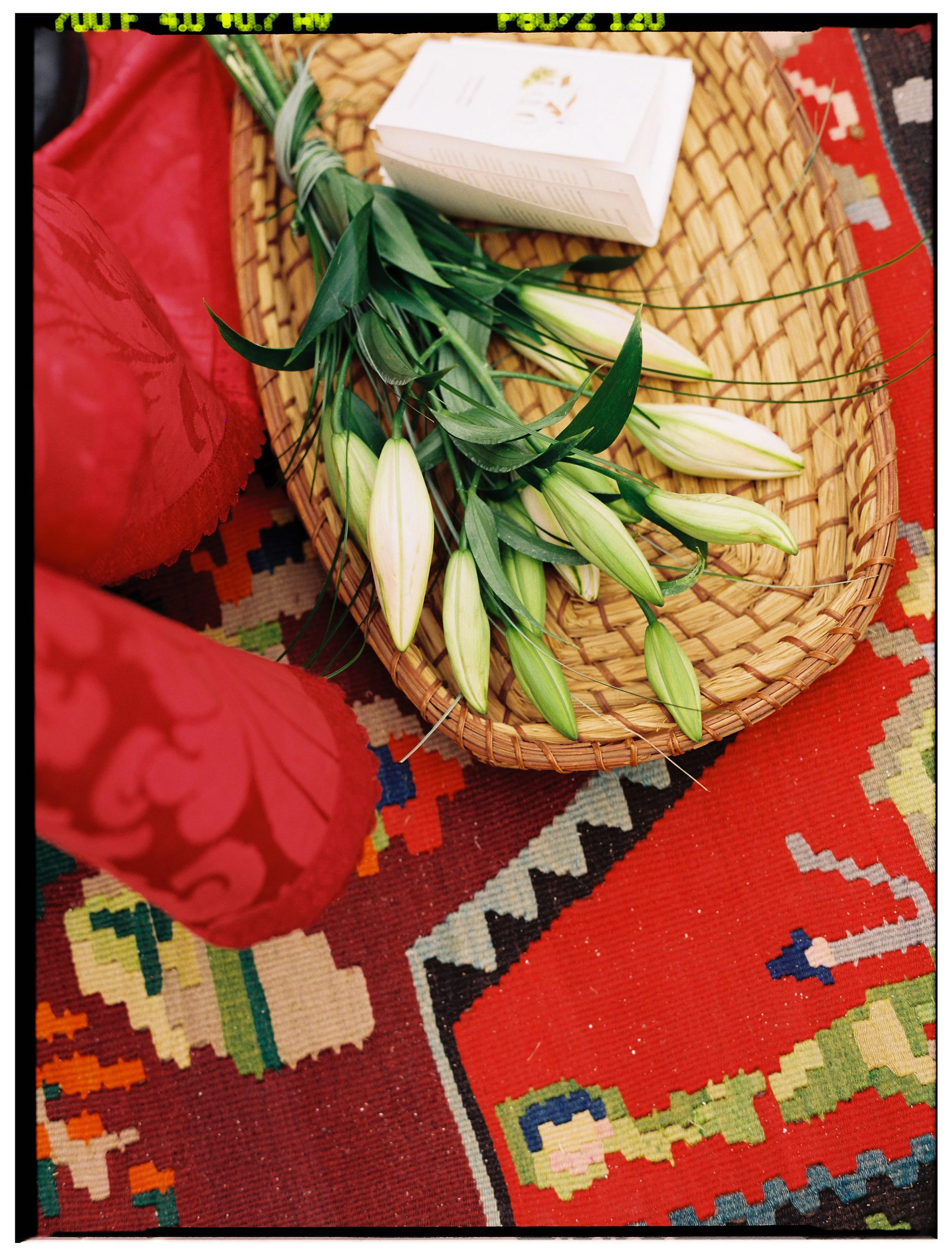 A woven basket with white lilies and green leaves next to a white box, placed on a colorful patterned rug with red, black, beige, blue, green, and orange elements.