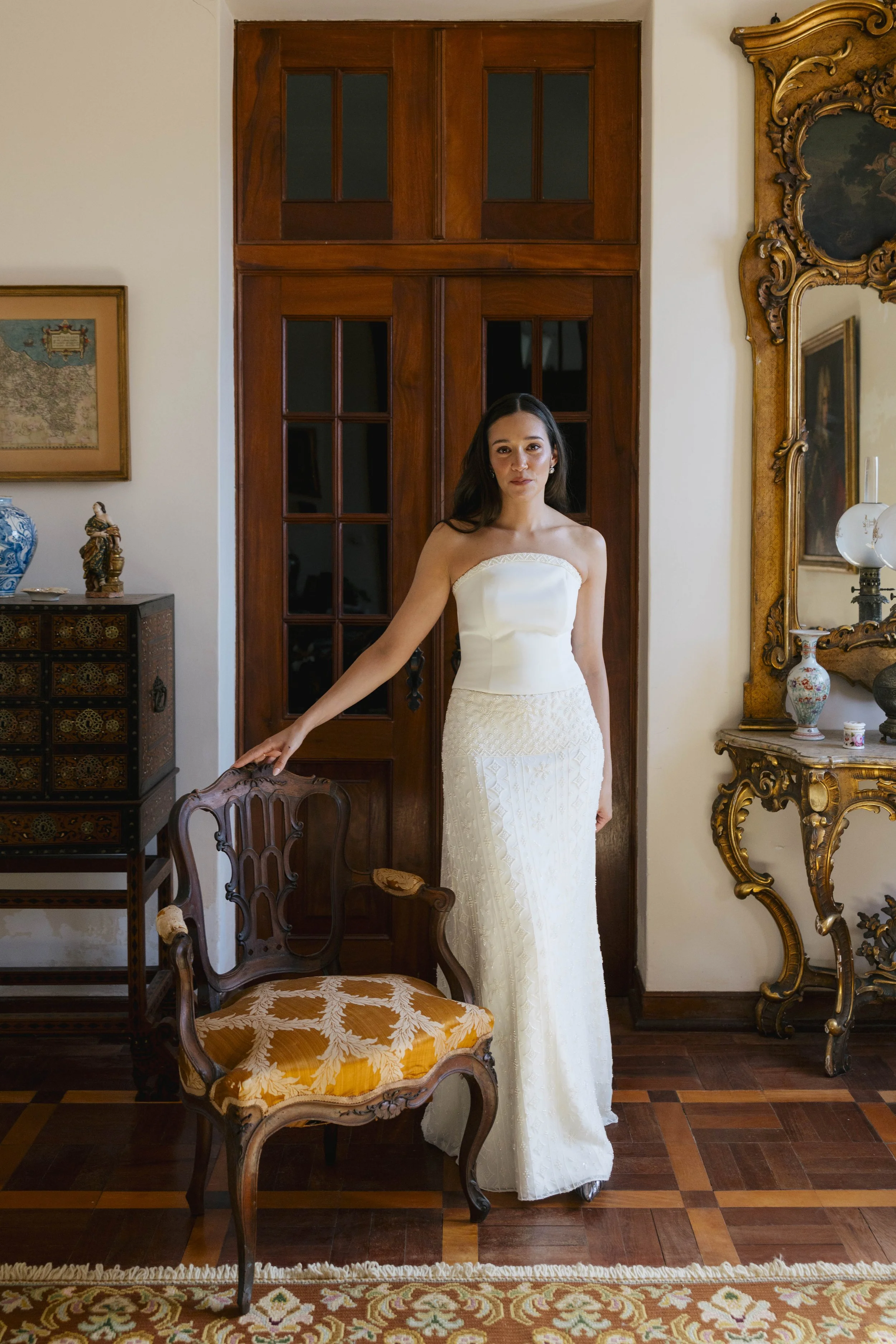 A woman in a strapless white wedding dress standing in a vintage room with wooden furniture, ornate mirror, and framed artwork.