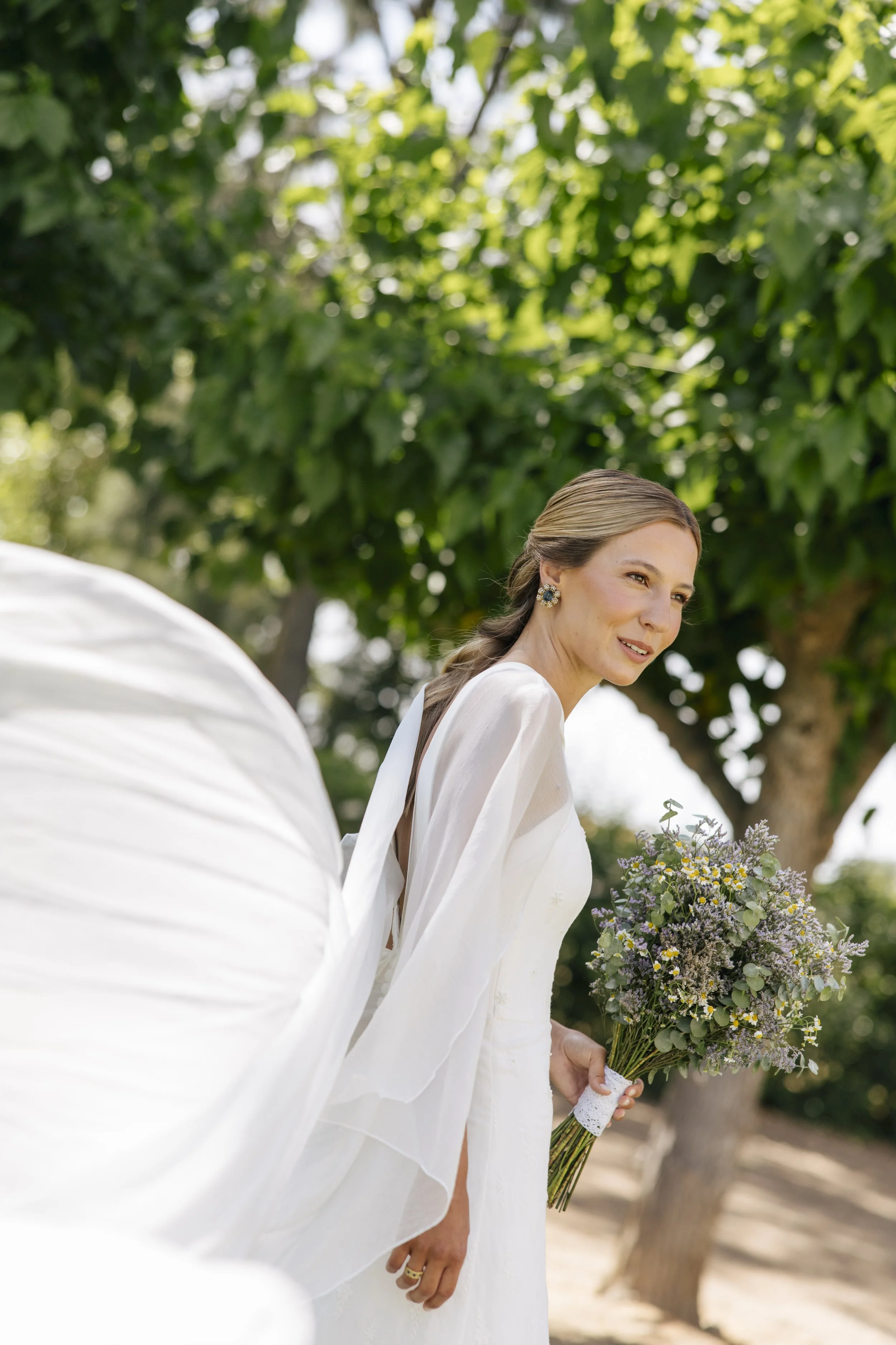 A woman dressed in white holding a bouquet of flowers outdoors with green trees in the background.